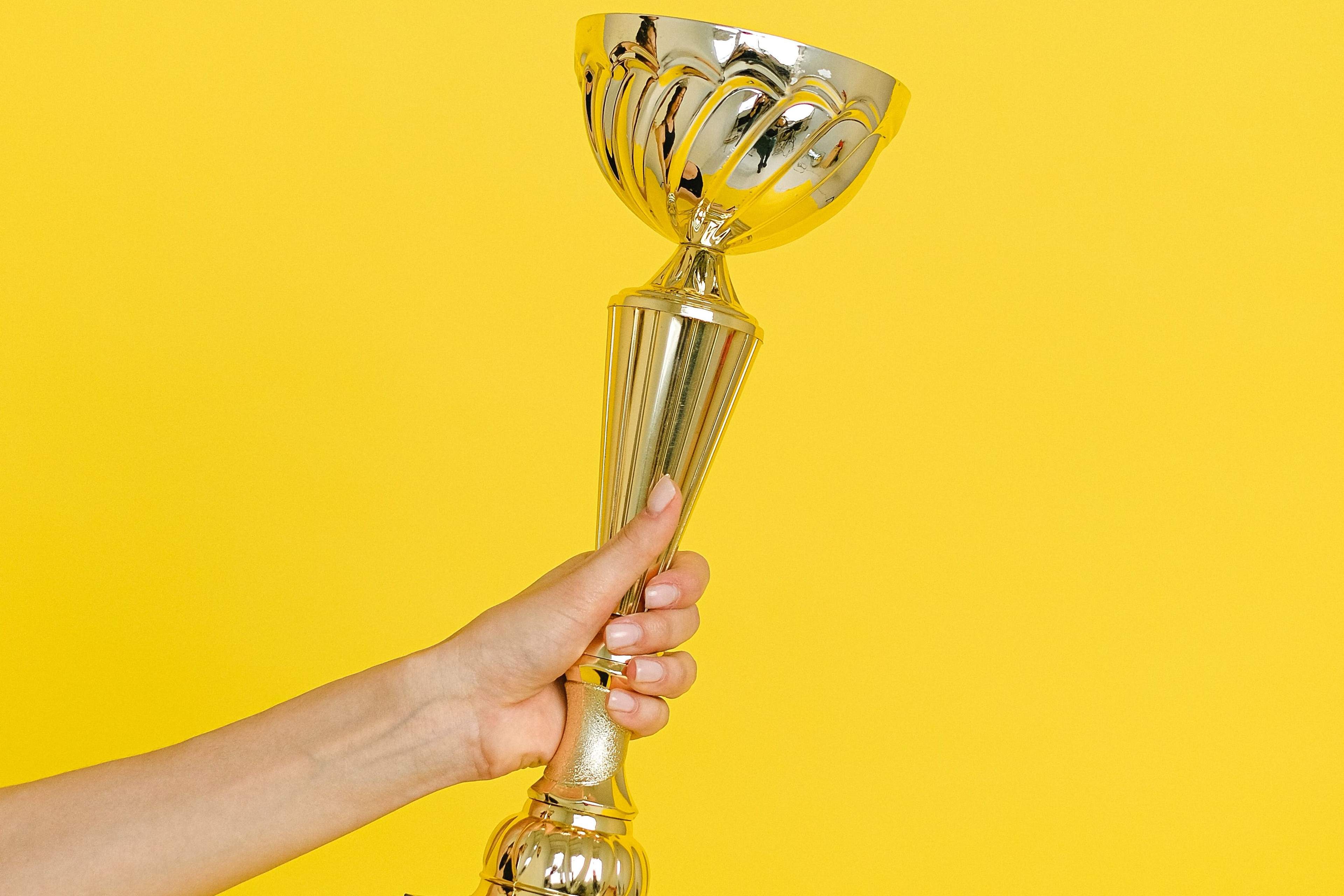 A bright yellow background. An arm reaches in from outside the frame, holding a large gold award.