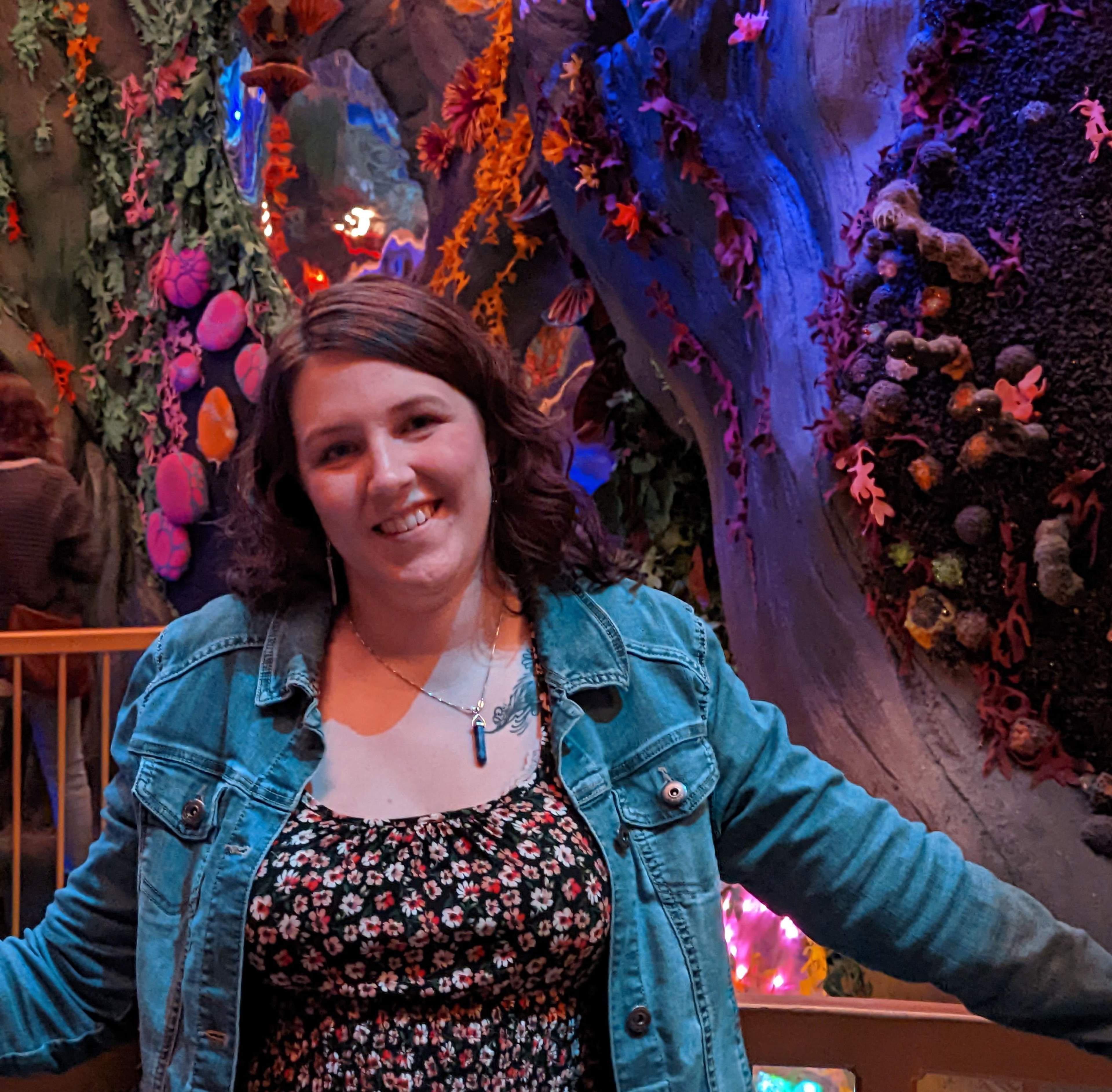 A photo of a white woman with long brown hair, blue eyes, wearing a big smile. She is standing in a fantasy woods art exhibit of felt trees