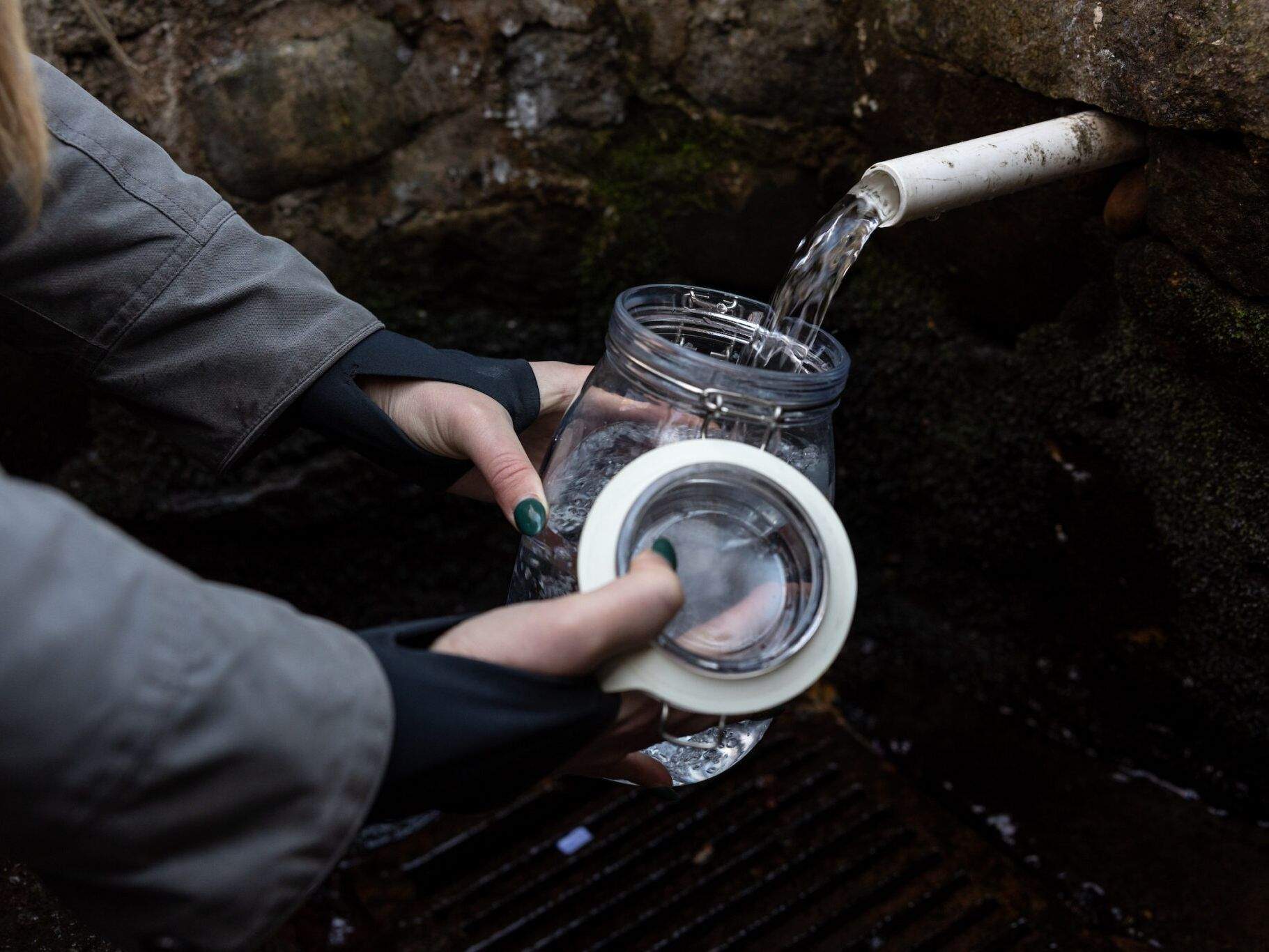 A woman's hand fills jars and bottles with water from a spring in the wall, she wears warm clothes to stay out of the rain.  