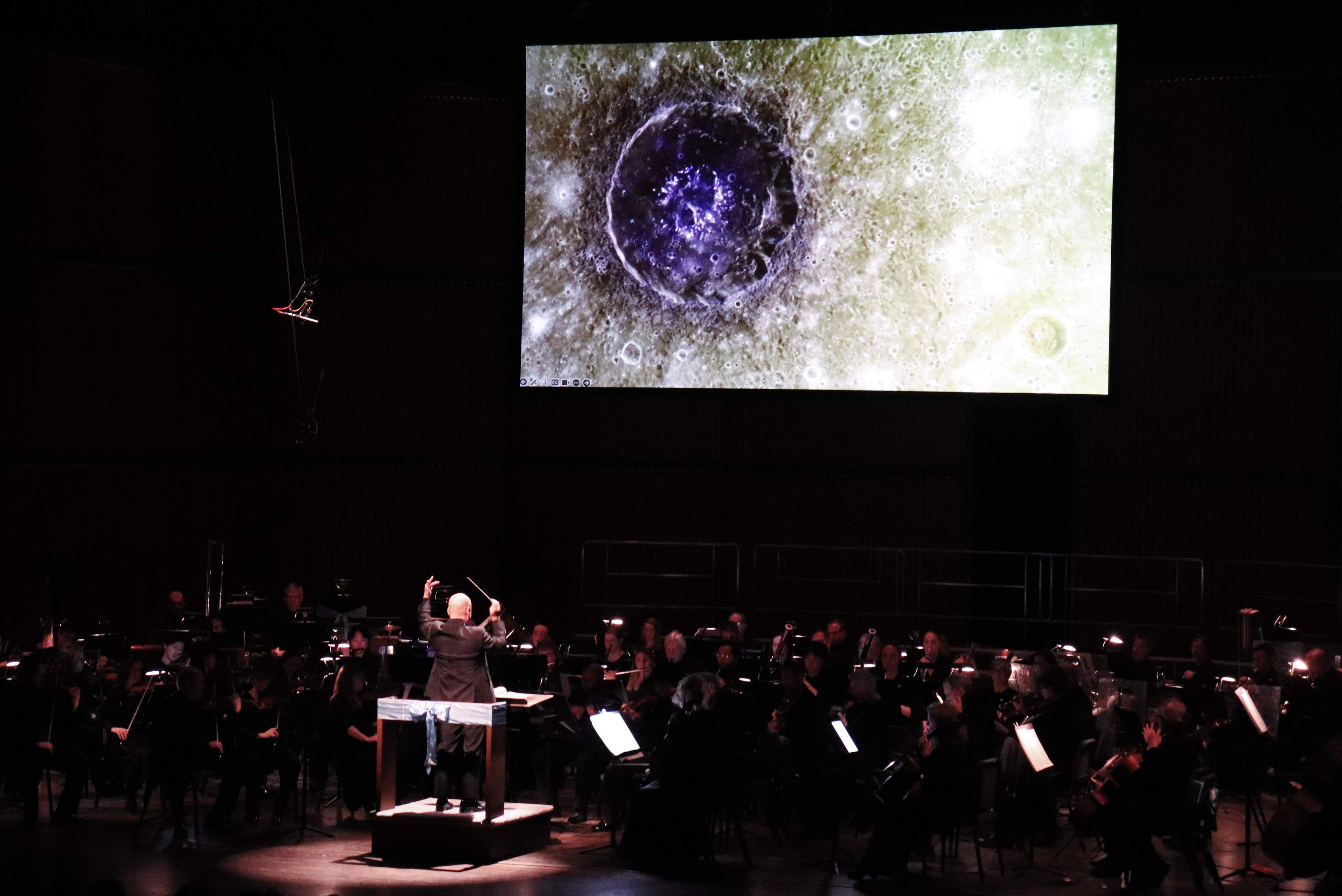 Symphony orchestra performing on a dimly lit stage with planets projected on screen