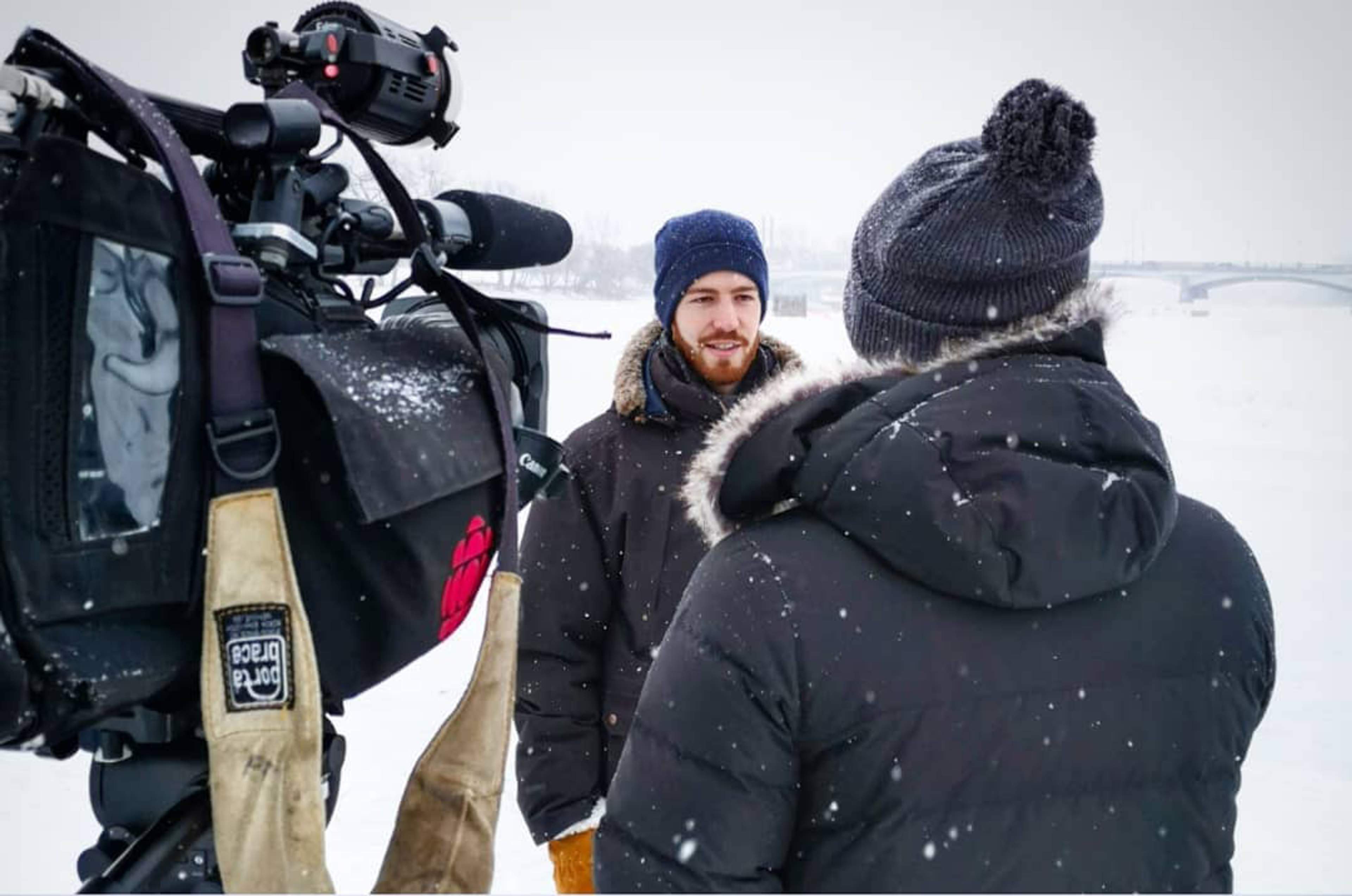 In a snowy landscape, Luke Rempel is interviewed by a CBC news reporter with a large camera. 