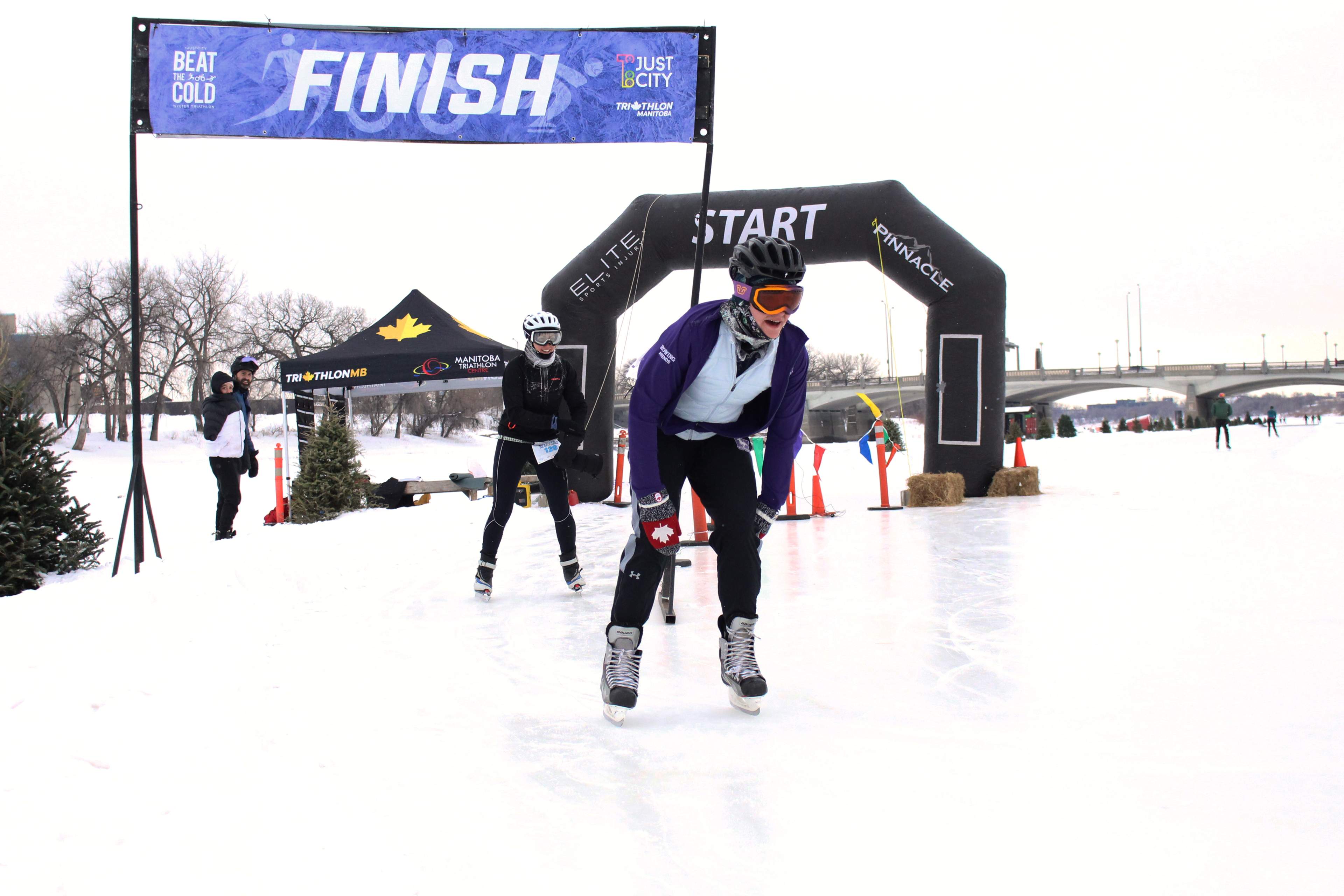 Two ice skaters wearing winter gear cross a finish line on ice. 