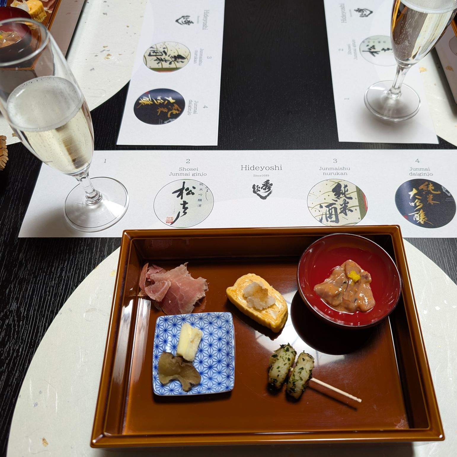 A sake tasting at Suzuki Brewing, Daisen, Akita — small bites on a lacquer tray alongside a Hideyoshi tasting card.