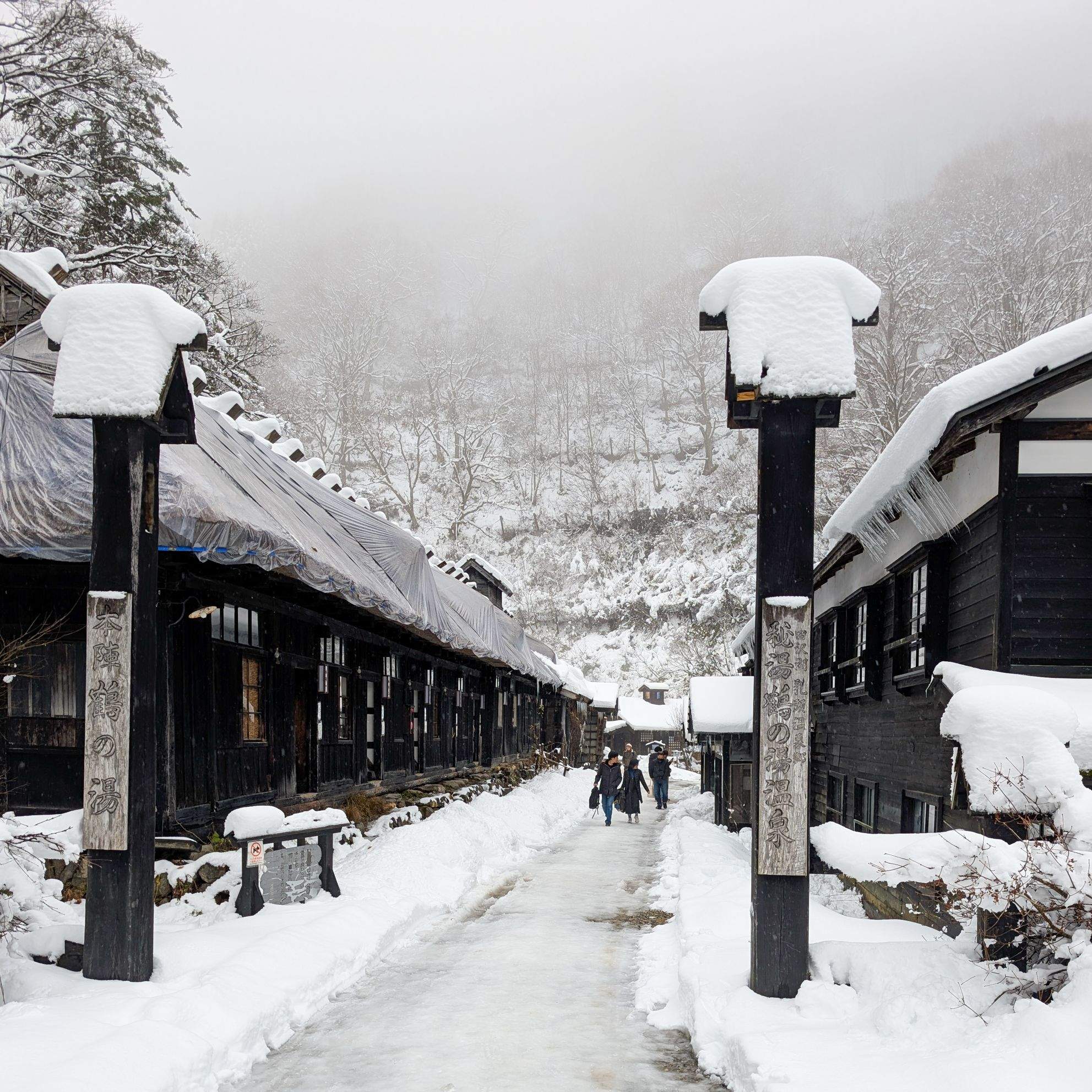 A snow-covered lane at Tsurunoyu Onsen, Nyuto Onsen Village, Akita, with traditional wooden buildings and visitors walking ahead.