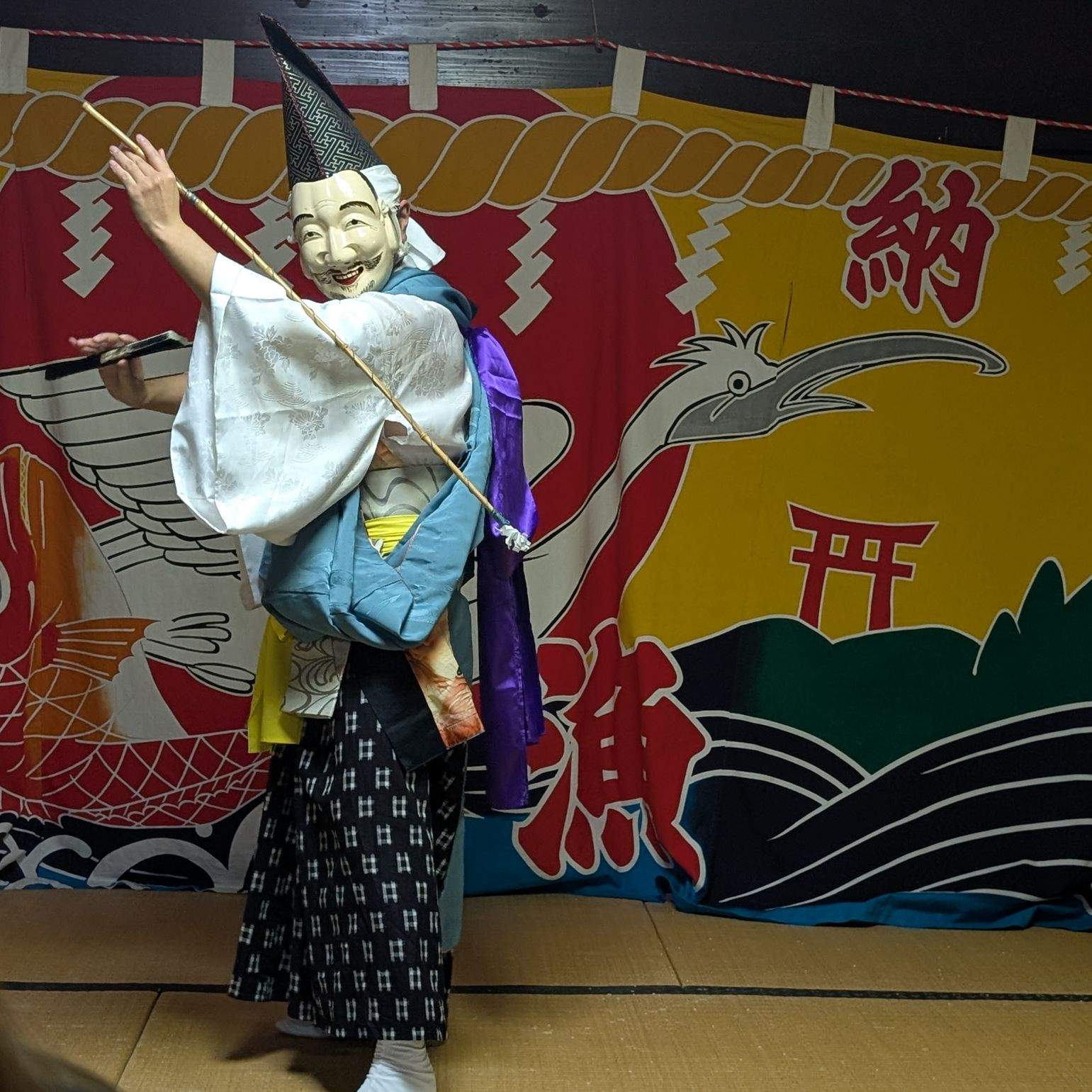 A masked Kagura performer in traditional costume holds a staff and fan on a tatami stage before a colorful festival backdrop, Iwate, Japan.