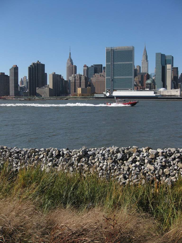 View of Manhattan skyline from Queens with a red boat speeding up the East River beneath a clear blue sky.