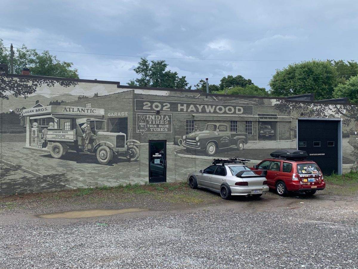 Cars parked outside of a garage with a monochrome mural on the side wall depicting the same building 75 years ago, Asheville, NC.
