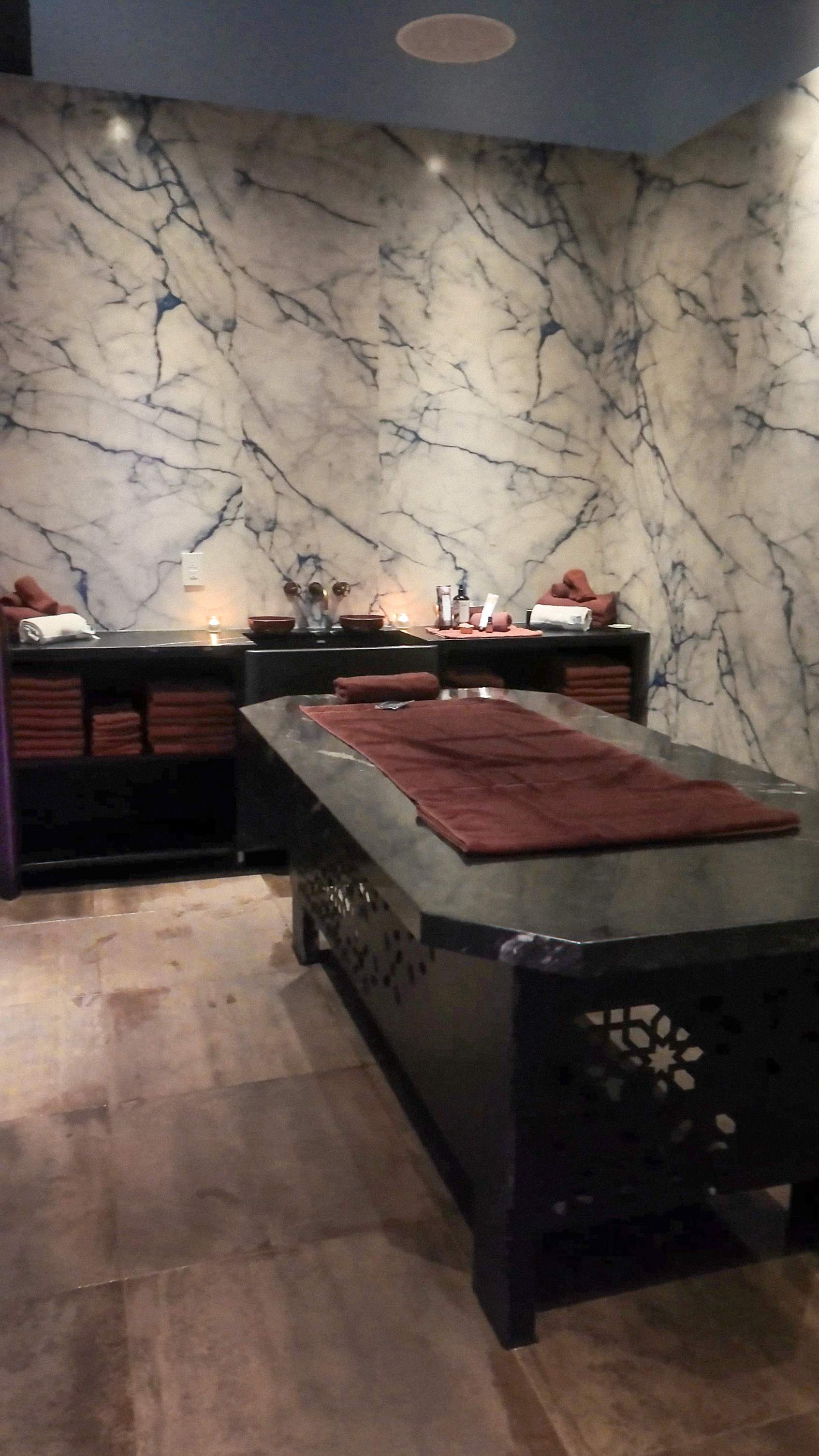 A luxurious Hammam Spa treatment room with dark marble-patterned walls, a massage table draped in brown towels, and subtle candle lighting f