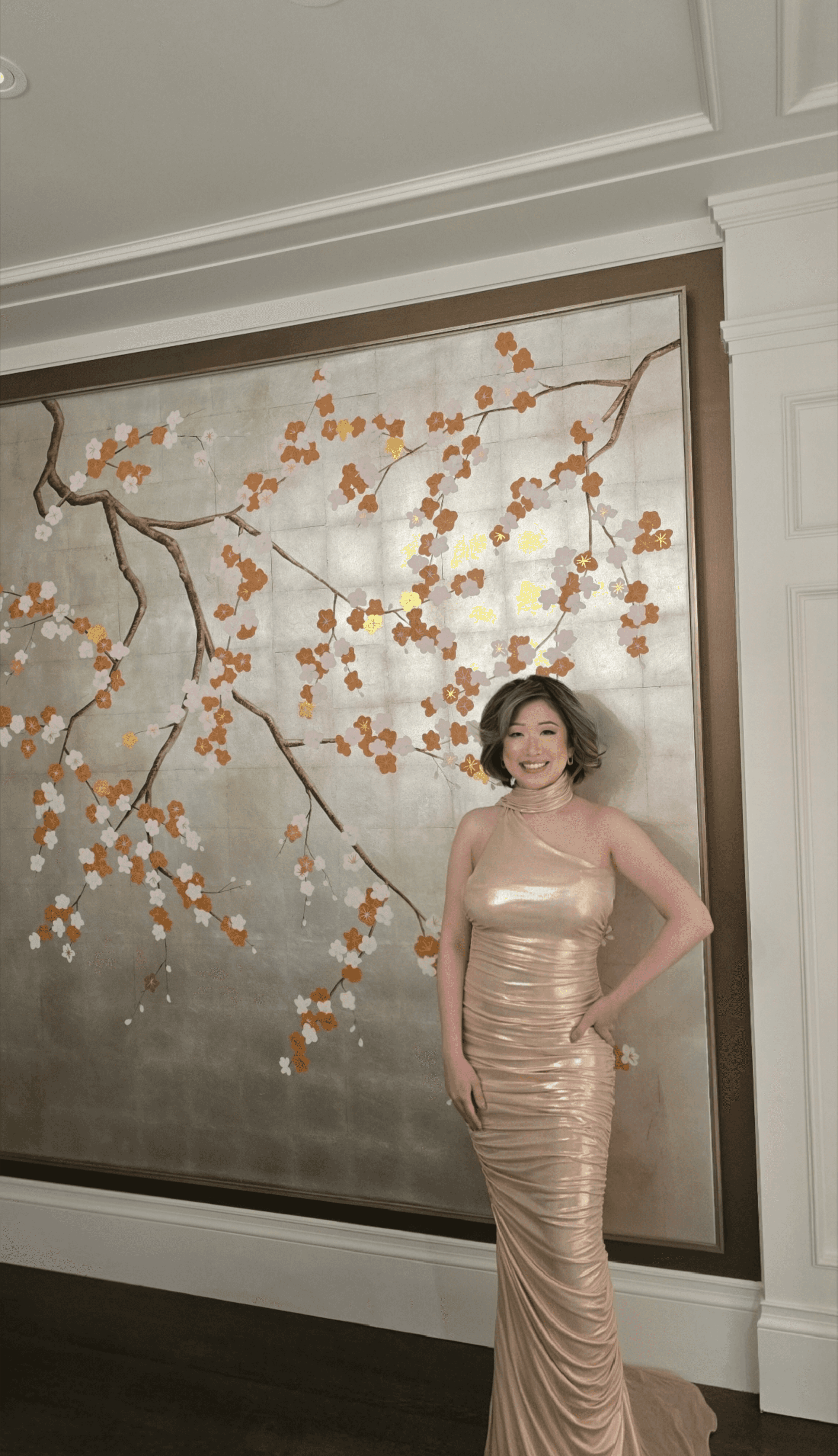 Betty Lam (@itsbettylam) smiling in a gold gown in front of a stunning, hand-painted de Gournay cherry blossom wall panel at 40 Golf Ave