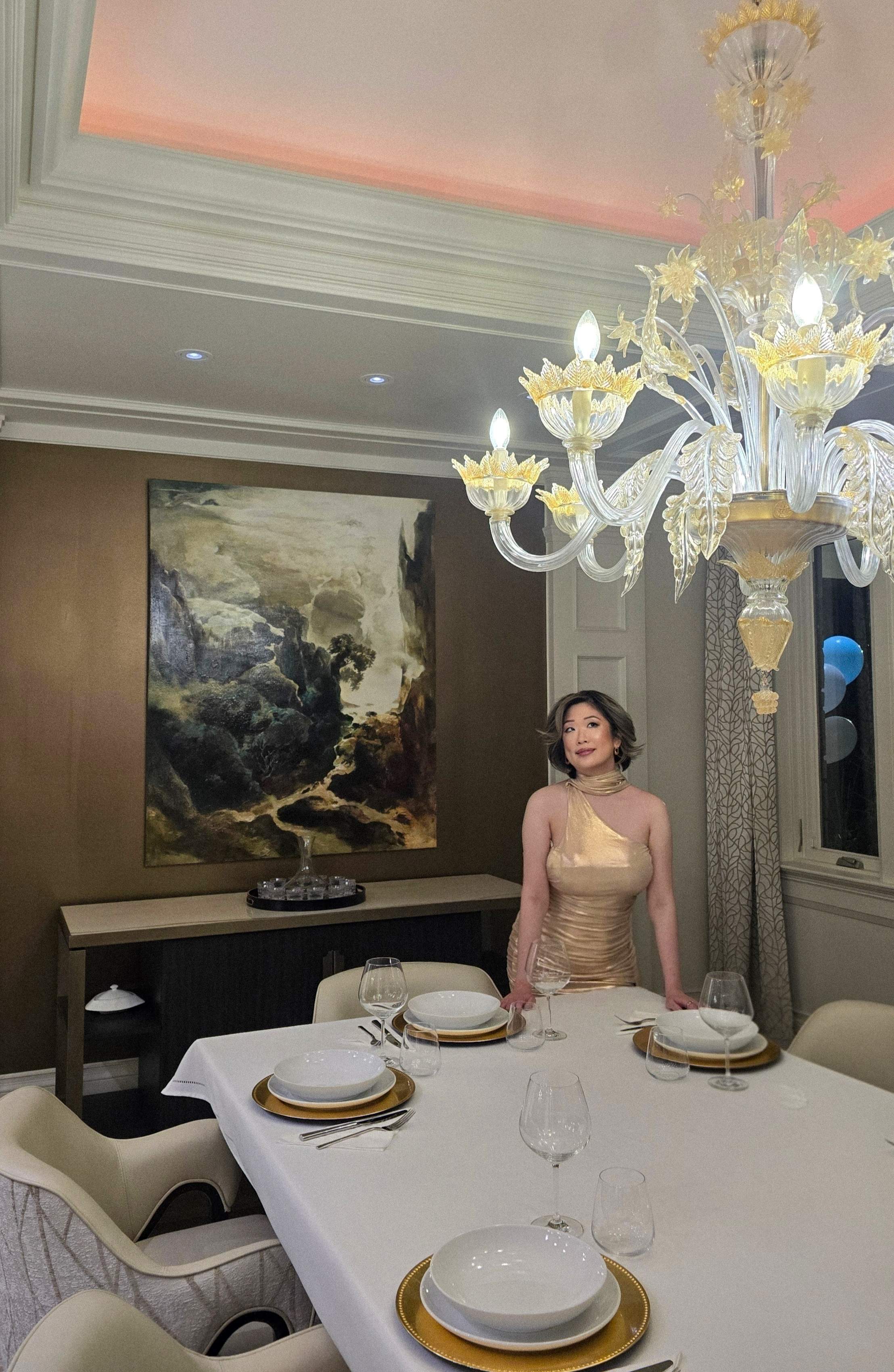 Betty Lam in the formal dining room of 40 Golf Avenue, standing by the grand Murano glass chandelier and formally set table.