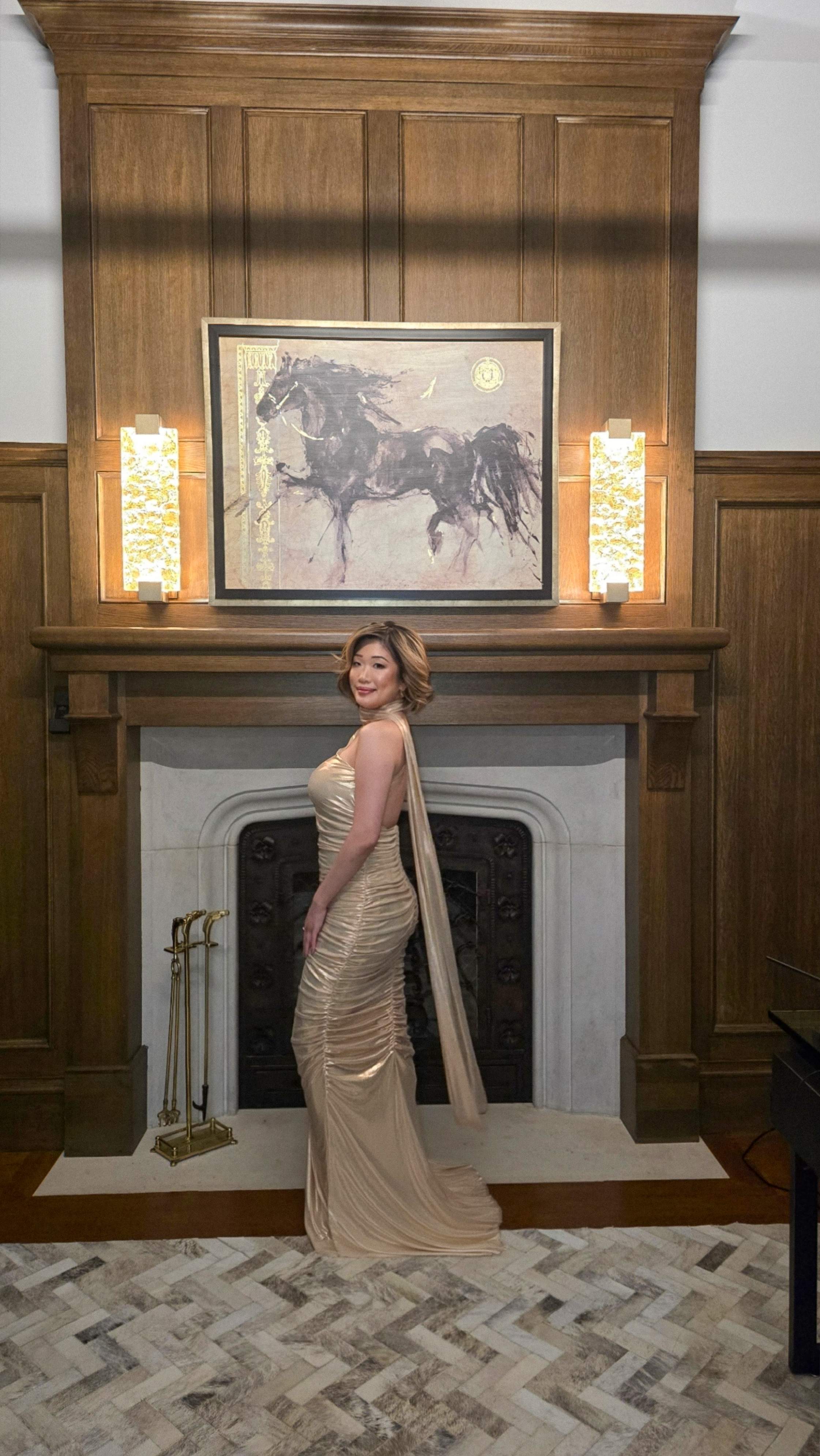 Betty Lam holds champagne in a gold gown, posing by the grand fireplace in the wood-paneled den of 40 Golf Avenue's luxury esta