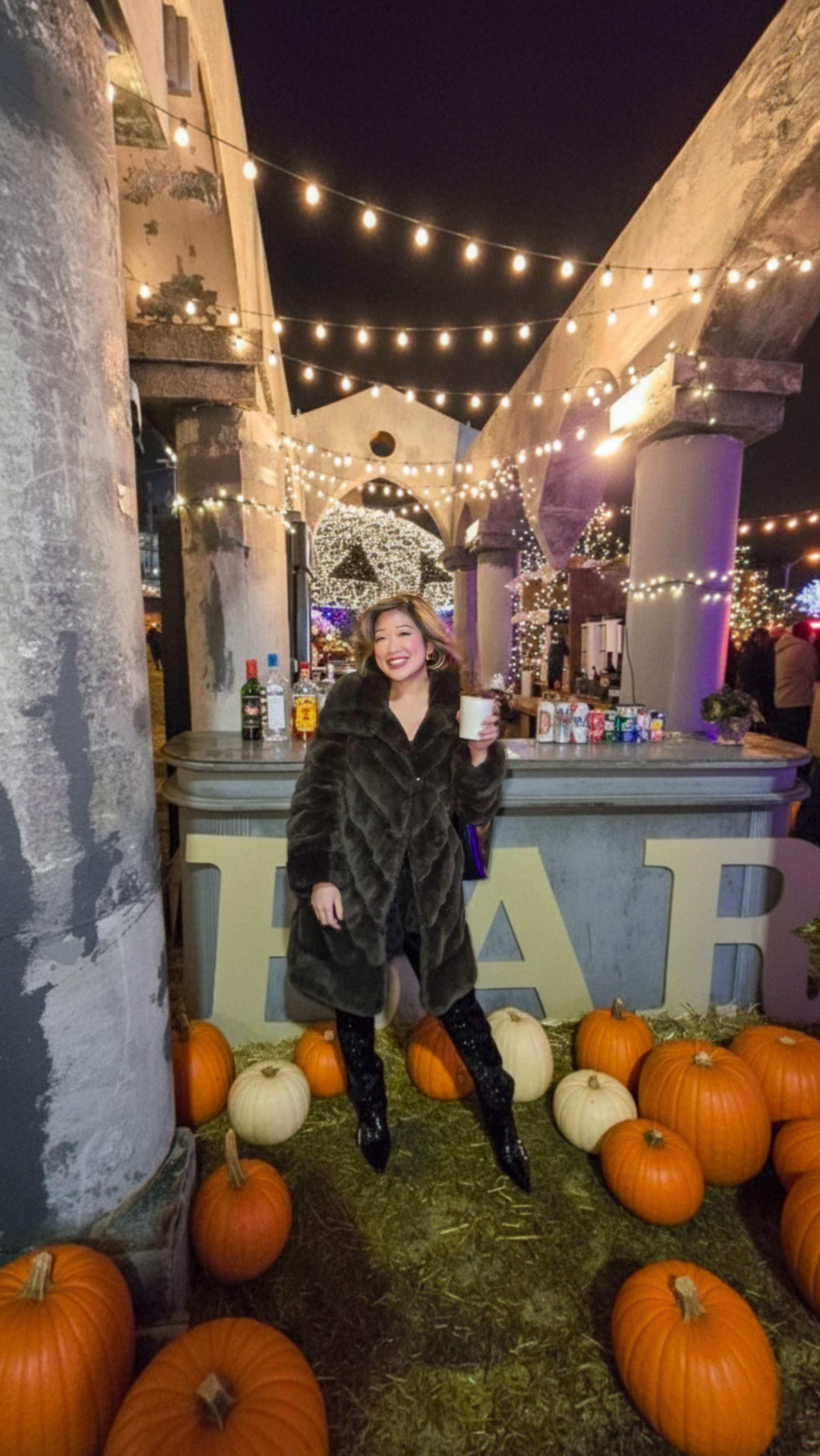 Betty Lam holding a drink at the Nights of Lights & Pumpkinville bar. Woman in fur coat at Toronto fall festival, smiling.