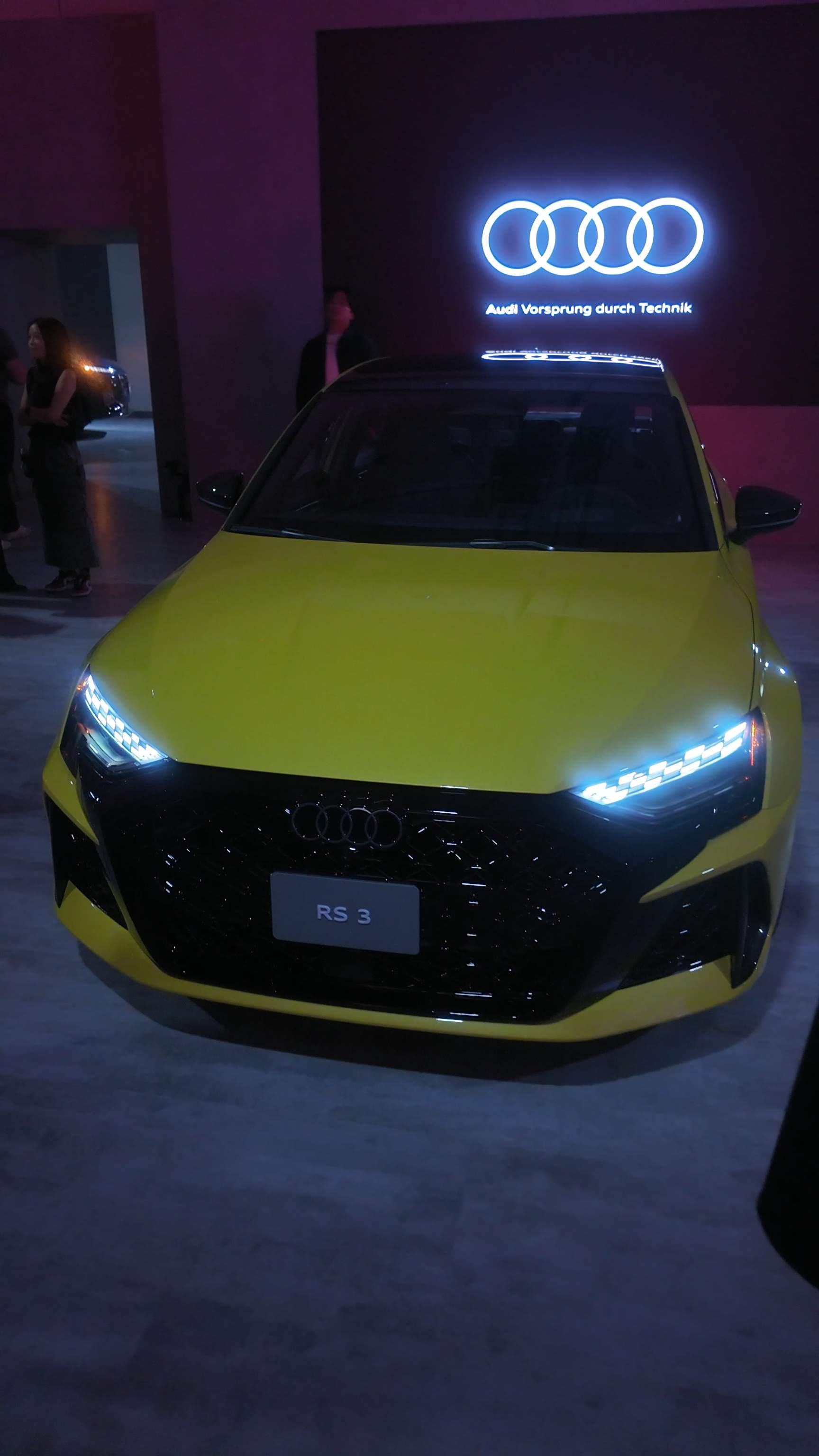 yellow Audi RS3 performance car with glowing daytime running lights parked on display beneath an illuminated Audi rings logo.