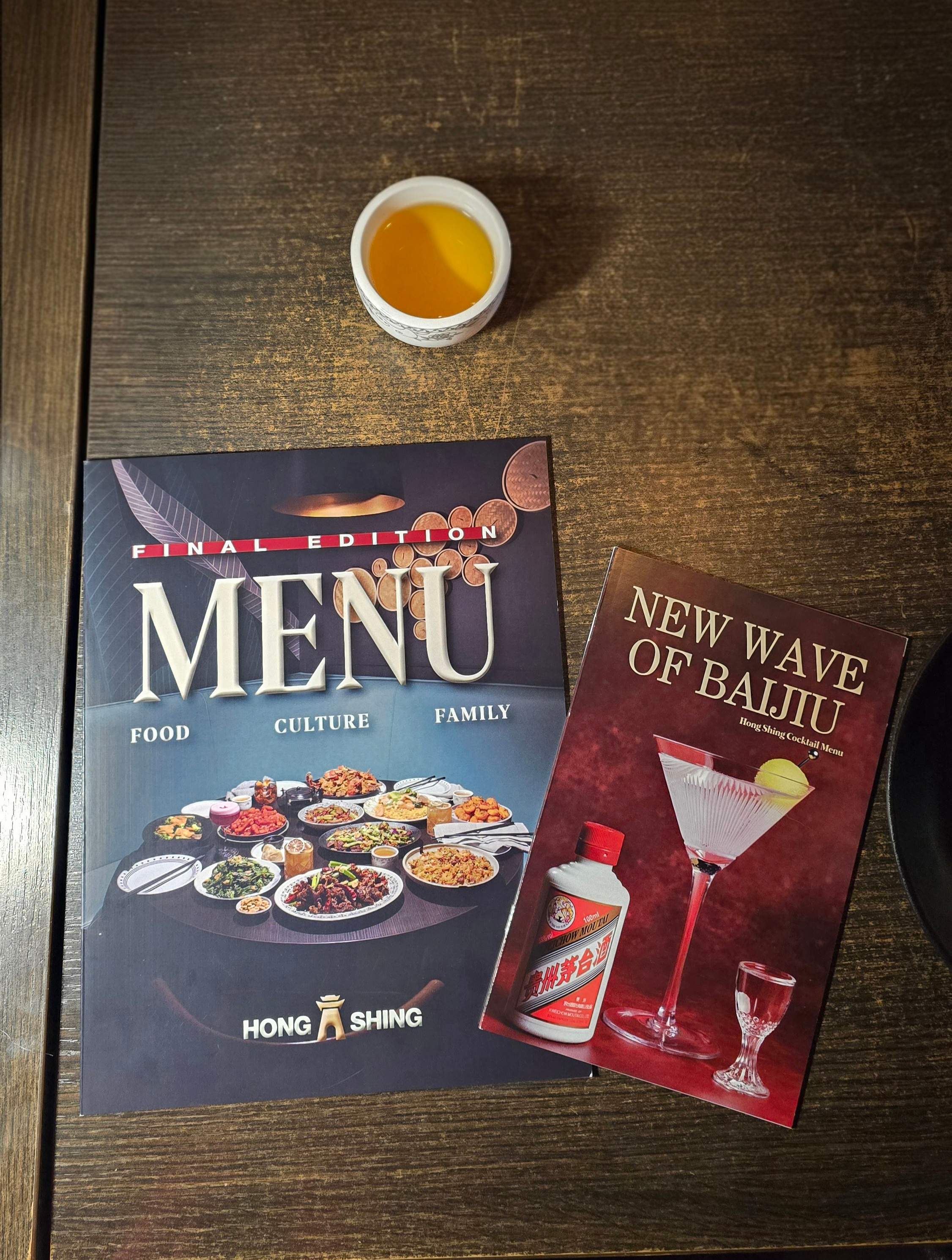 Hong Shing Toronto dining menu and New Wave of Baijiu cocktail menu on a wooden restaurant table next to a traditional cup of hot tea.