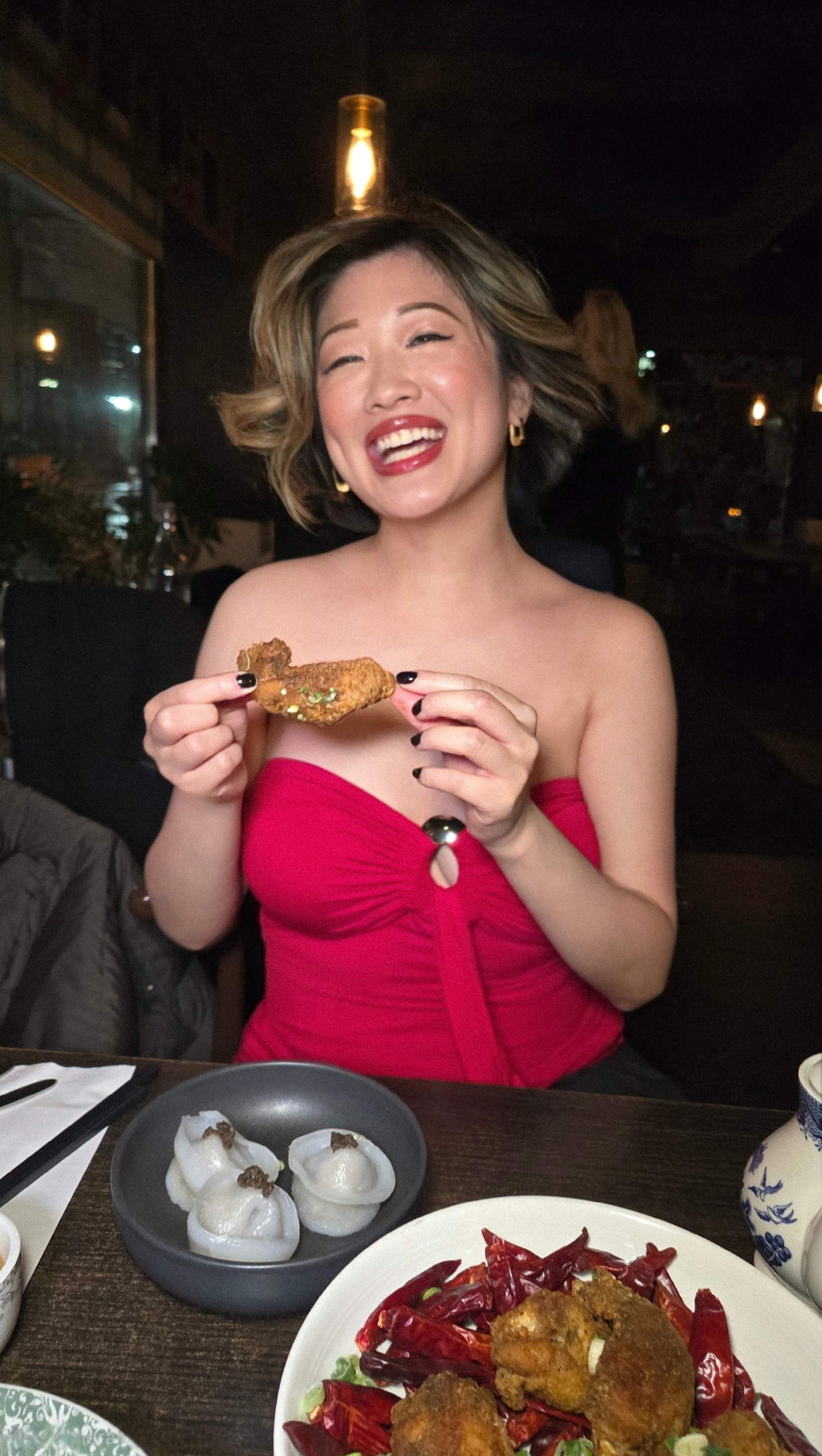 Woman smiling while holding a crispy Mala chicken wing at Hong Shing restaurant Toronto, surrounded by a Halal Chinese food feast.
