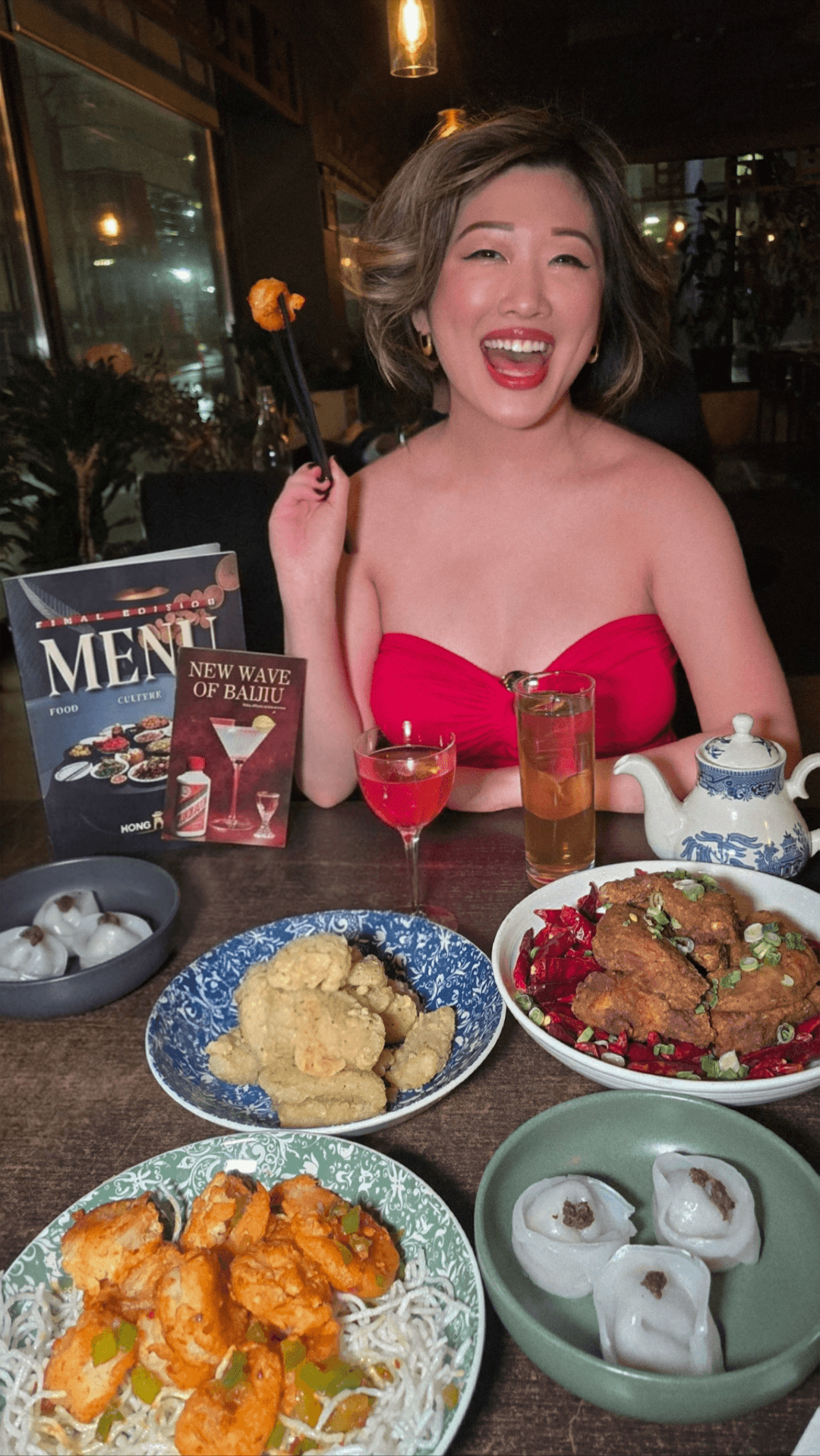 Smiling woman enjoying Halal Chinese food and Baijiu cocktails at Hong Shing in downtown Toronto. Table full of spicy shrimp and dumplings.
