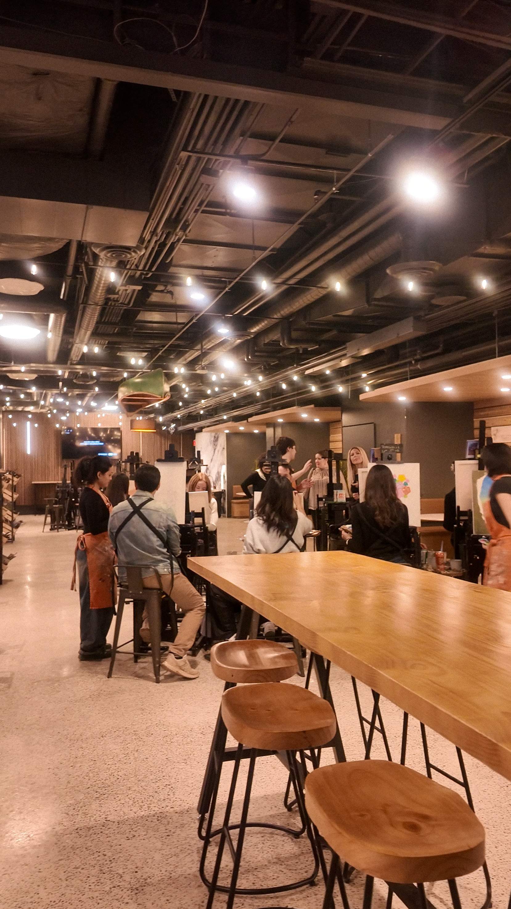 Guests painting at easels inside the spacious Paint Cabin studio, featuring long wooden tables, string lights, and a relaxed atmosphere. 