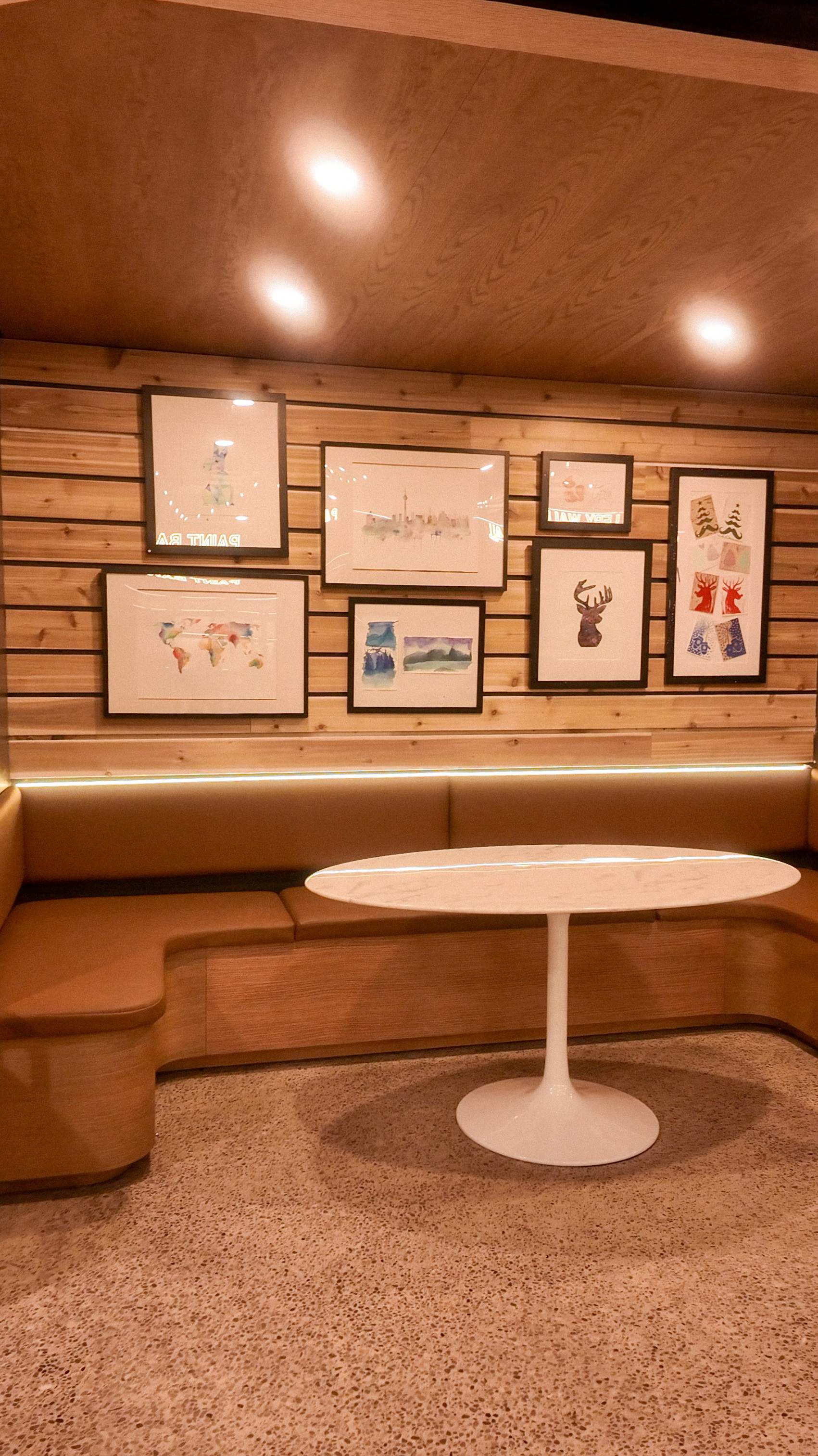 Cozy seating area at Paint Cabin featuring a leather banquette, modern white table, and framed watercolor art on wood paneled walls