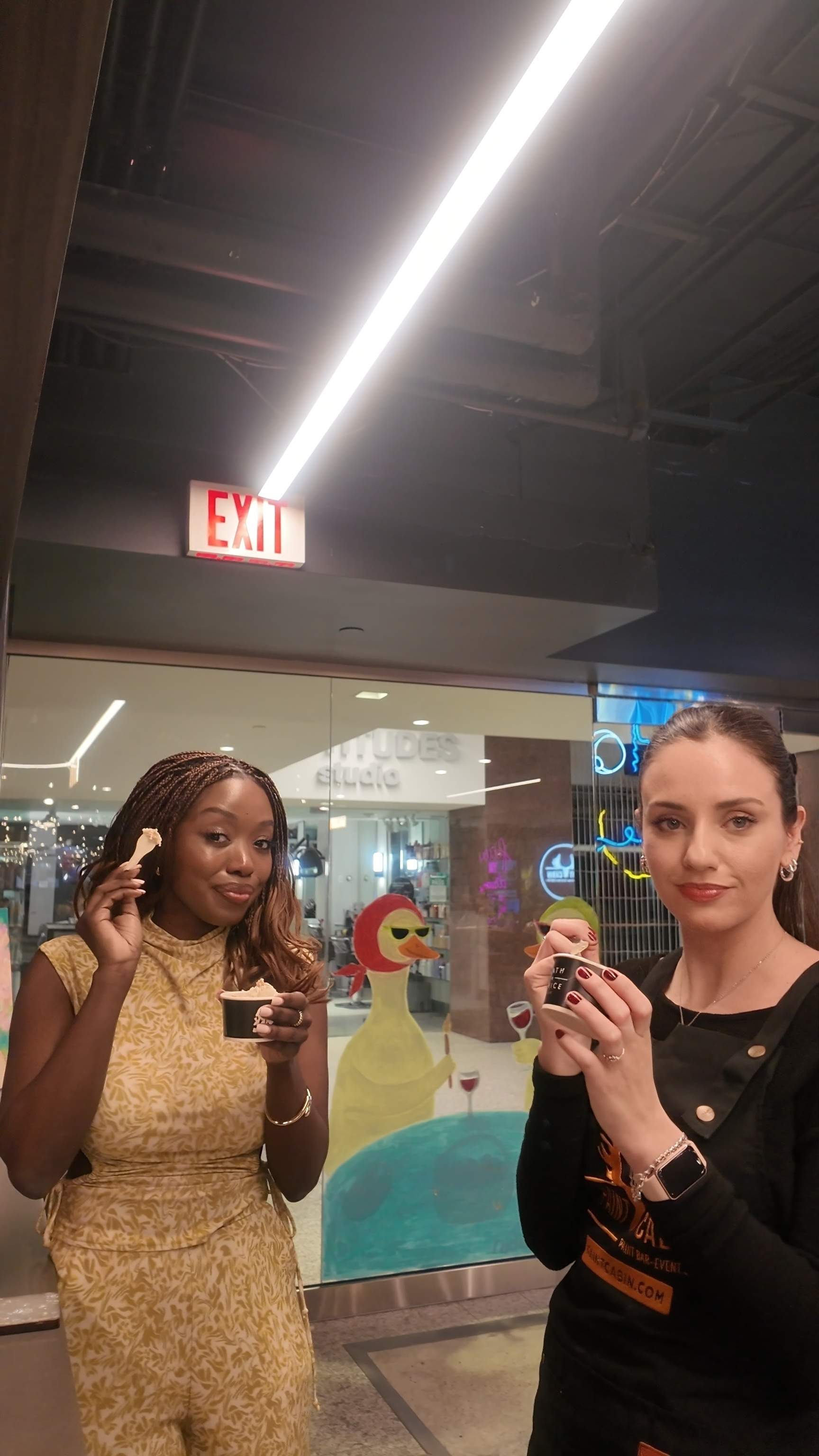 Two women enjoying cups of gelato after an evening of painting, standing near the glass entryway of Paint Cabin in Scotia Plaza