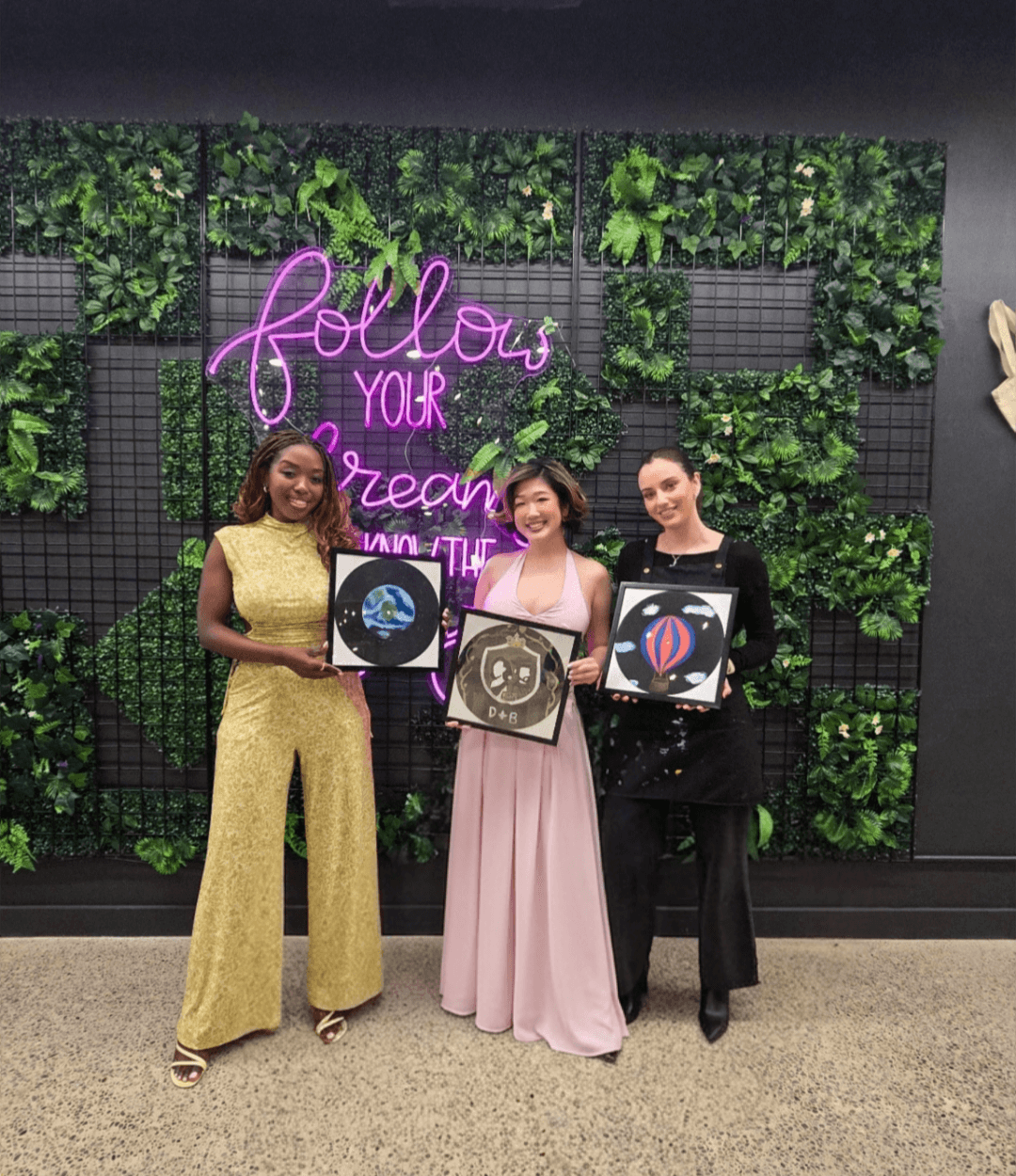 Three women holding finished vinyl record paintings, standing in front of a botanical wall with a pink neon sign at Paint Cabin.