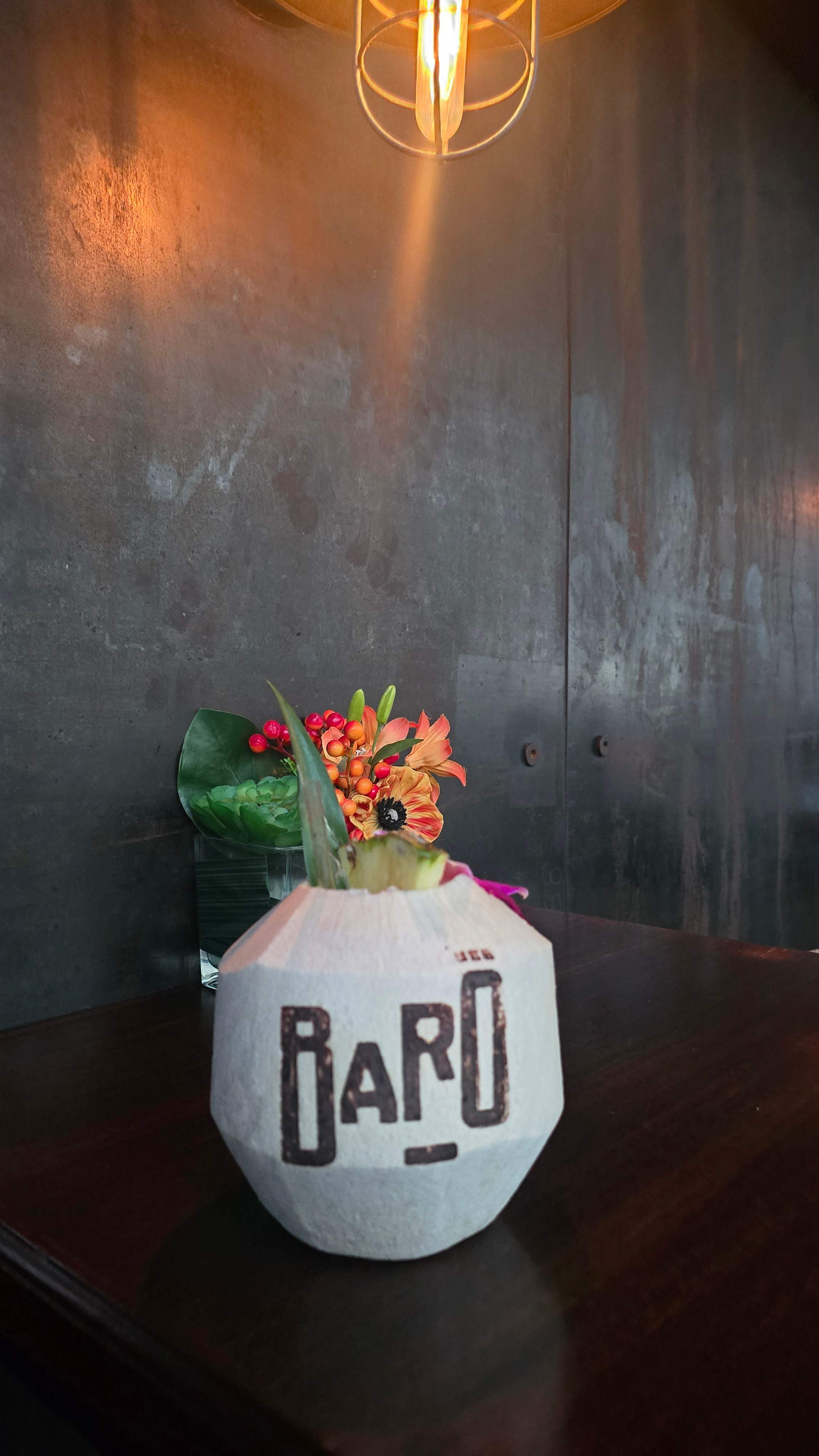 Whole white coconut branded with the Baro logo, served as a tropical cocktail with orchid and pineapple garnishes