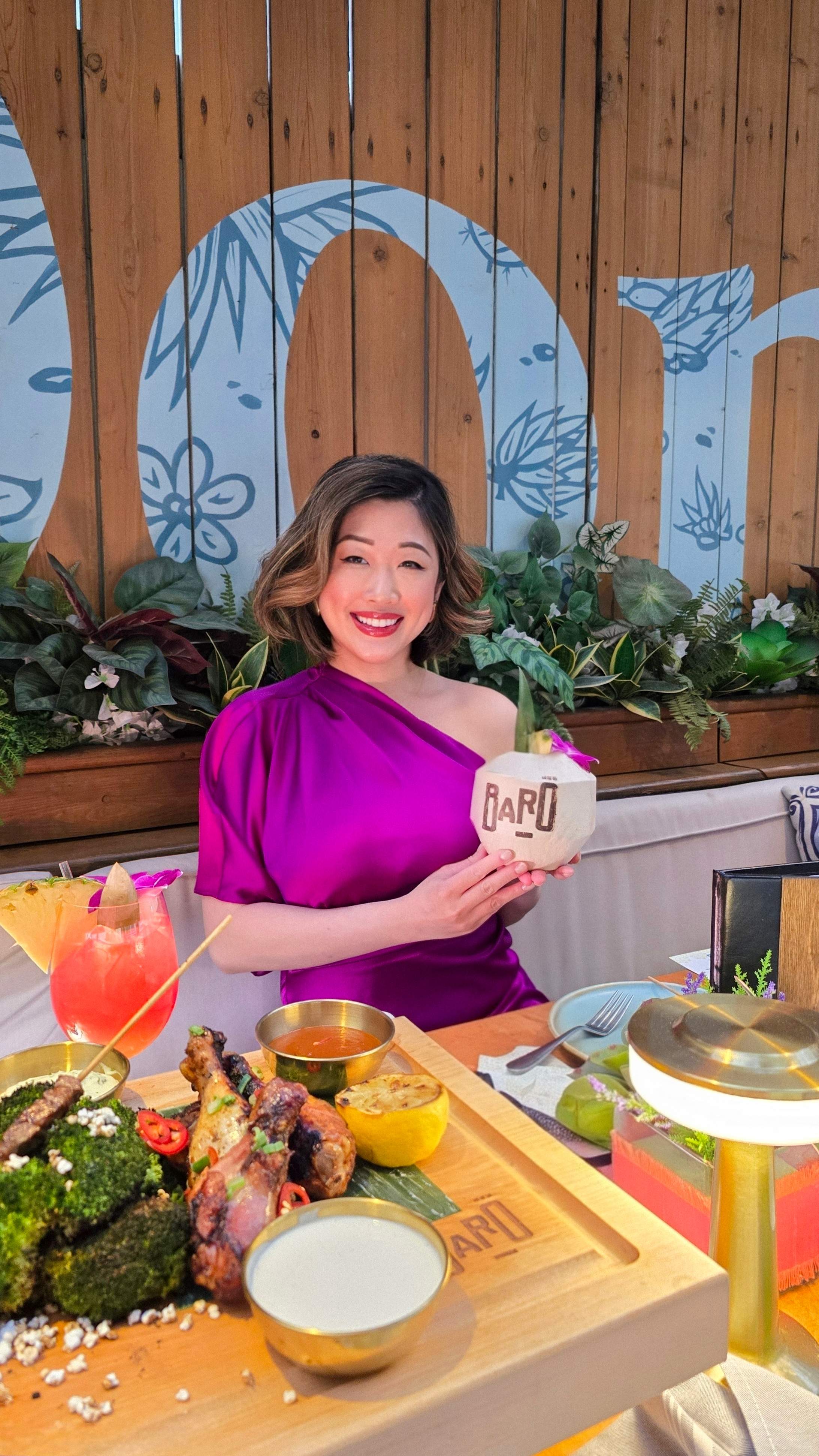 Close-up of smiling woman in purple dress holding a branded Baro coconut cocktail, with a detailed close-up of a food board. 