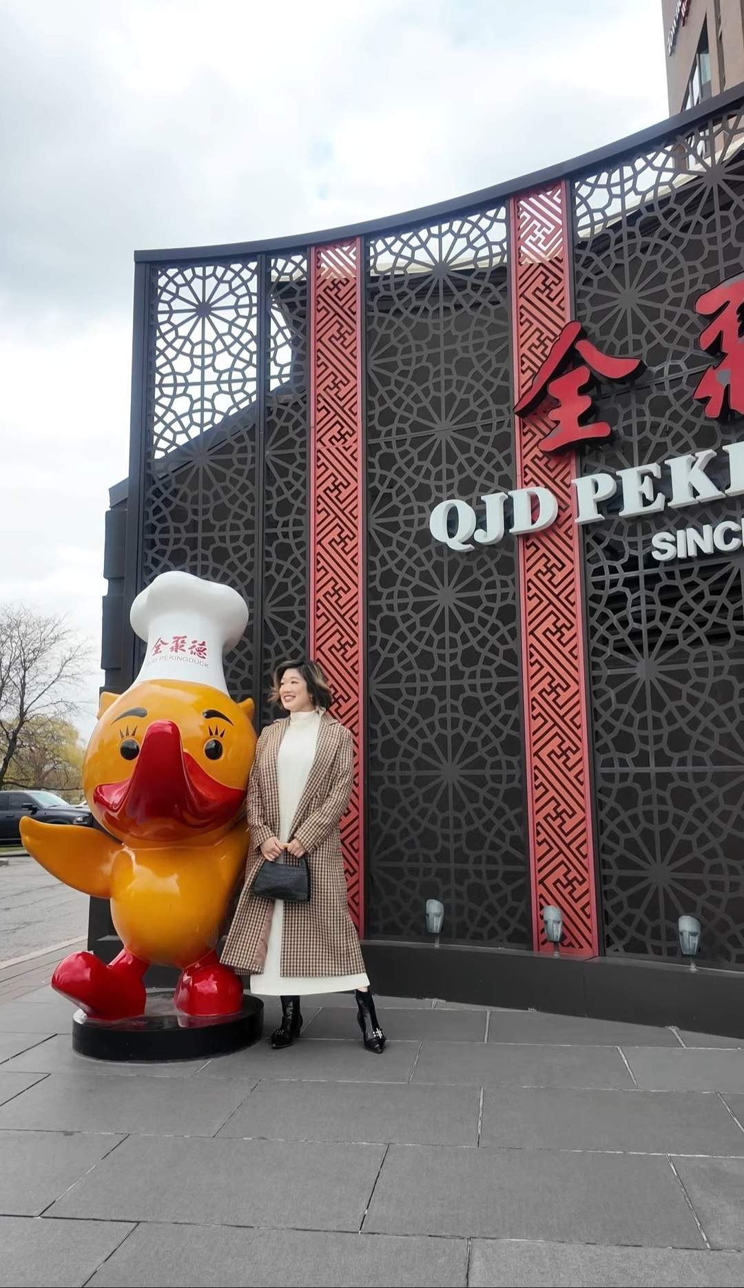 Woman posing and smiling beside QJD Peking Duck restaurant cartoon duck mascot statue outside Markham Ontario location on Woodbine Avenue QJ