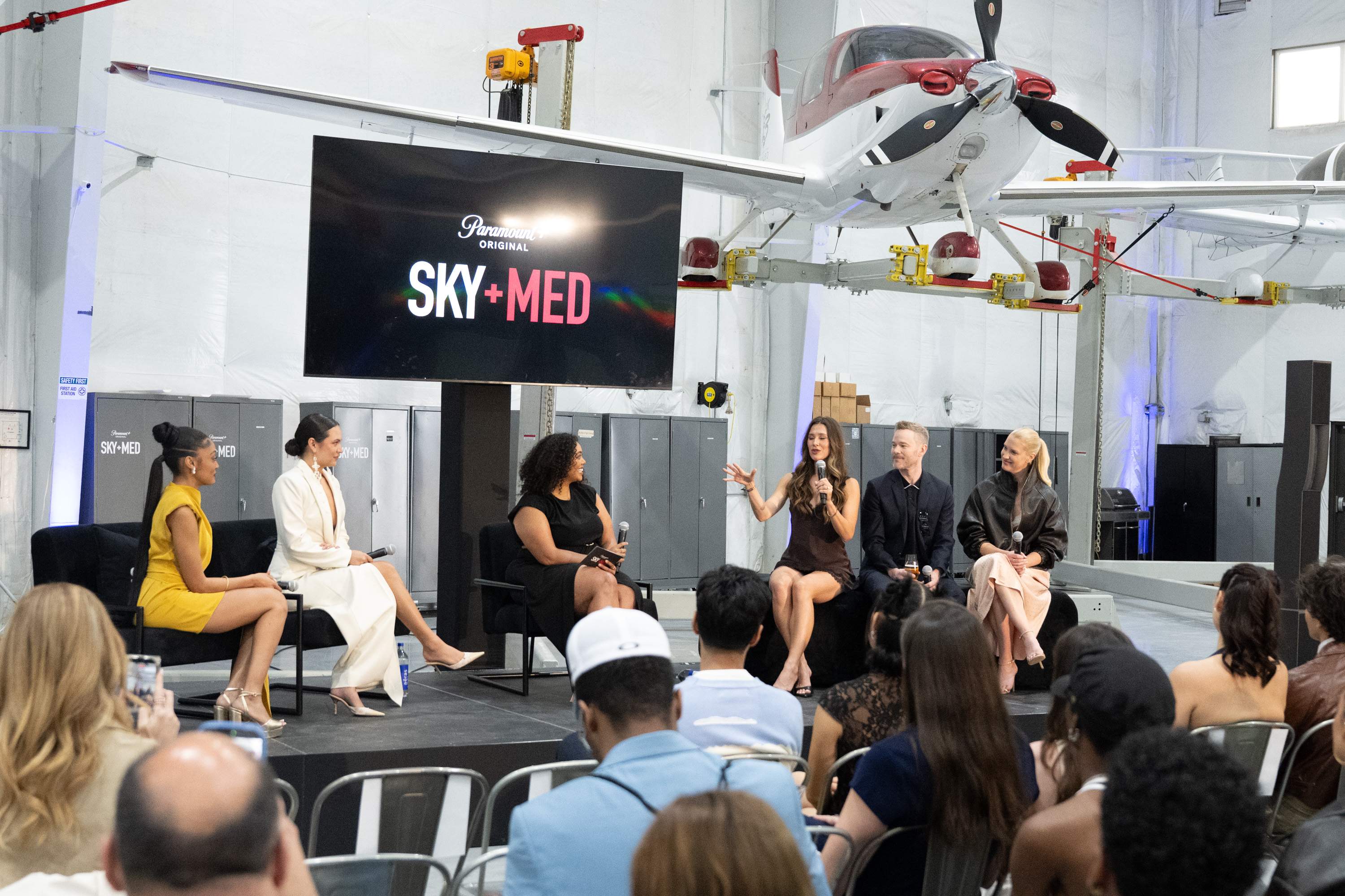 Wide view of the SkyMed Season 4 cast Q&A panel hosted by Brittnee Blair inside an airport hangar. The actors are seated on a stage below a 