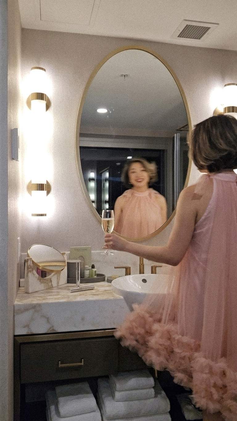 Woman in a pink tulle dress smiling and taking a mirror selfie with champagne in a luxury bathroom at The Sutton Place Hotel Halifax.