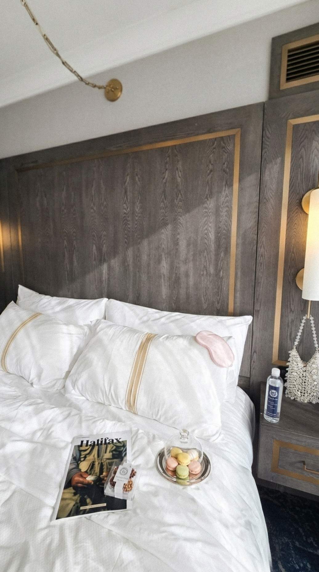 Luxurious white bedding at Sutton Place Hotel Halifax with a sleep mask, macarons, magazine, and bottled water on the nightstand.
