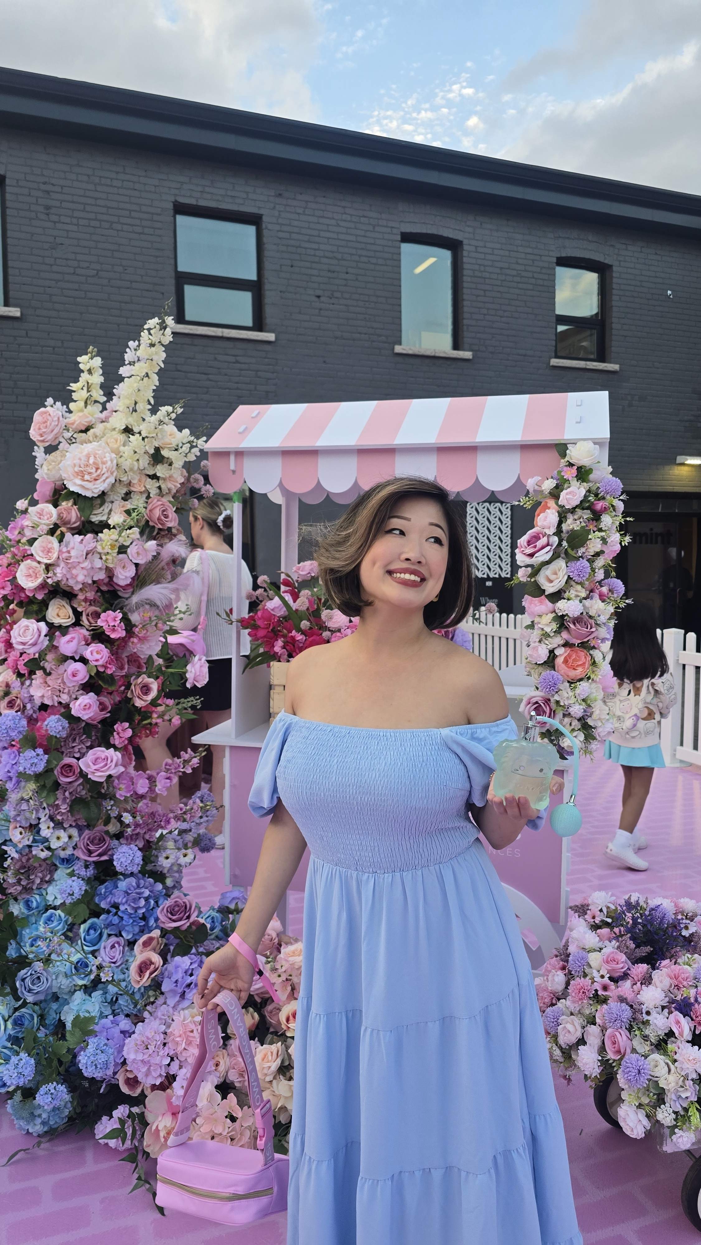 Girl posing with Squishmallows fragrance blue bottle. Candy striped Sephora and Squishmallows Fragrances market stall with a pastel flowers