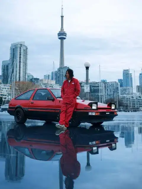 Sung Kang in a red racing jumpsuit leaning against a red Toyota AE86, with Toronto's CN Tower and skyline reflected on the wet ground behind