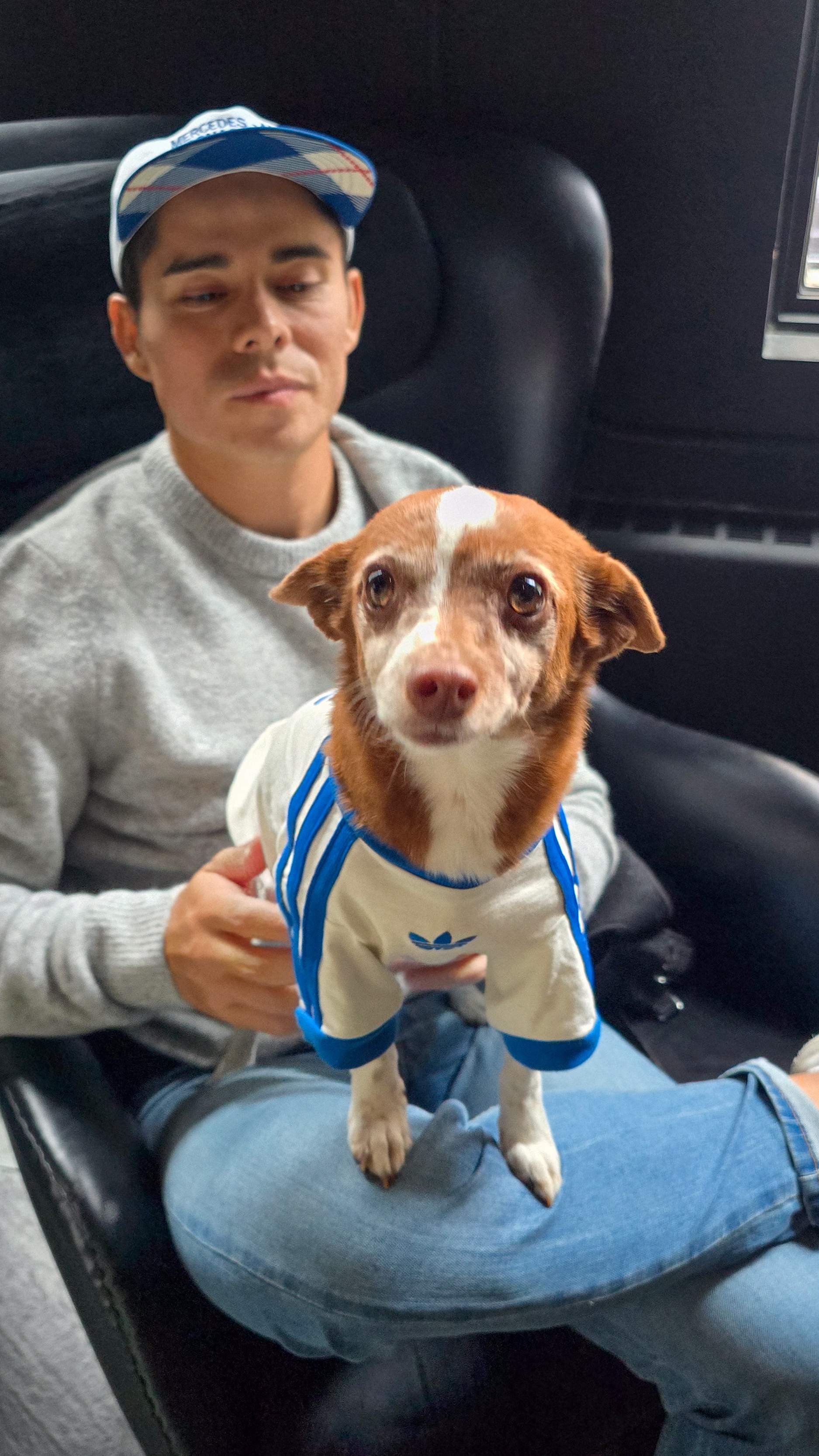 Man in a plaid Mercedes cap holding a small dog dressed in a white and blue Adidas-style tracksuit, sitting on a black leather chair