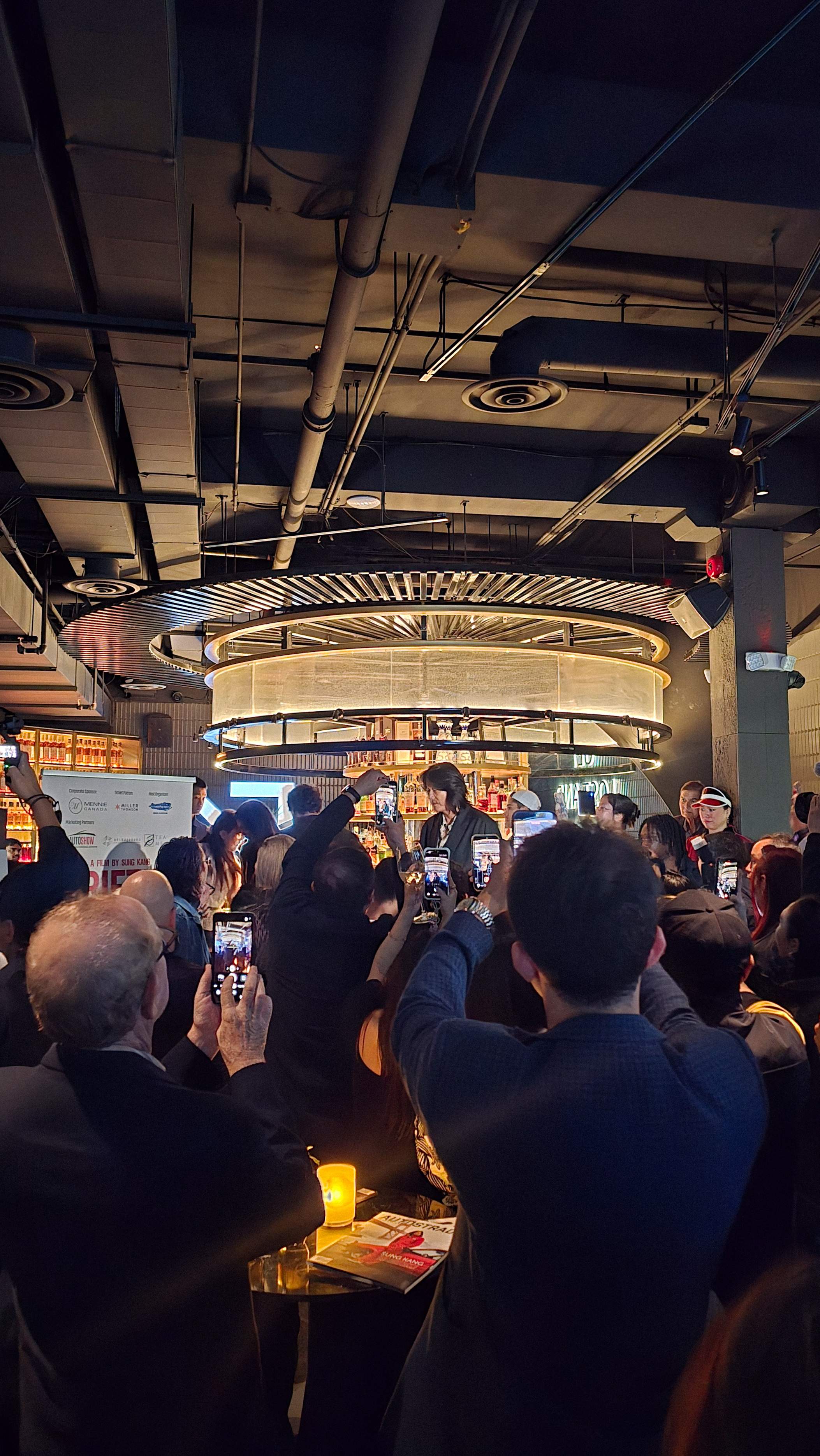 Crowd holding up phones to photograph Sung Kang near the bar at the DRIFTER reception, with a circular illuminated bar structure overhead 