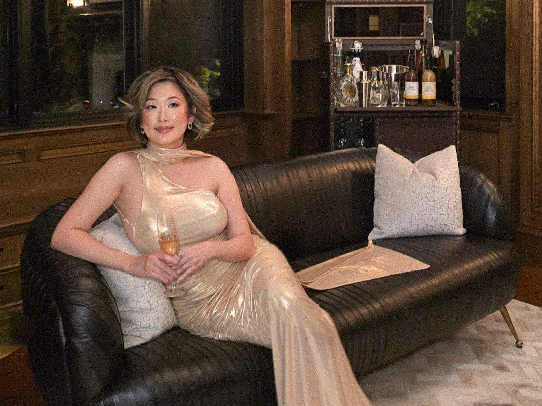 Betty Lam in a gold gown, holding champagne and lounging on a chic black leather sofa in the den of 40 Golf Avenue's luxury estate
