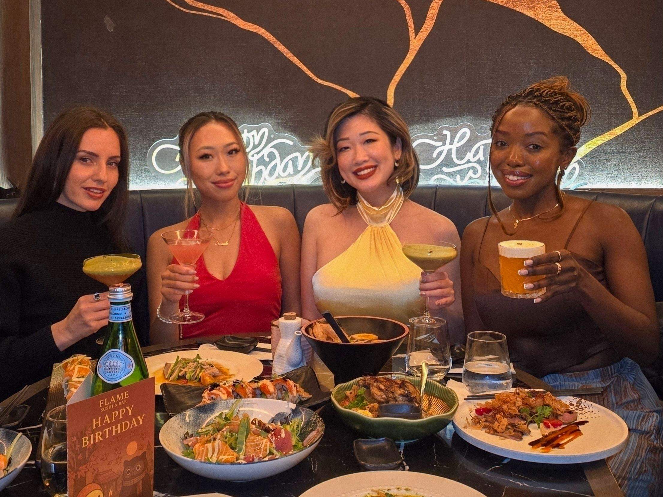 Four stylish women smiling and toasting with cocktails around a lavish table spread at Flame Sushi for a celebratory girls' night.