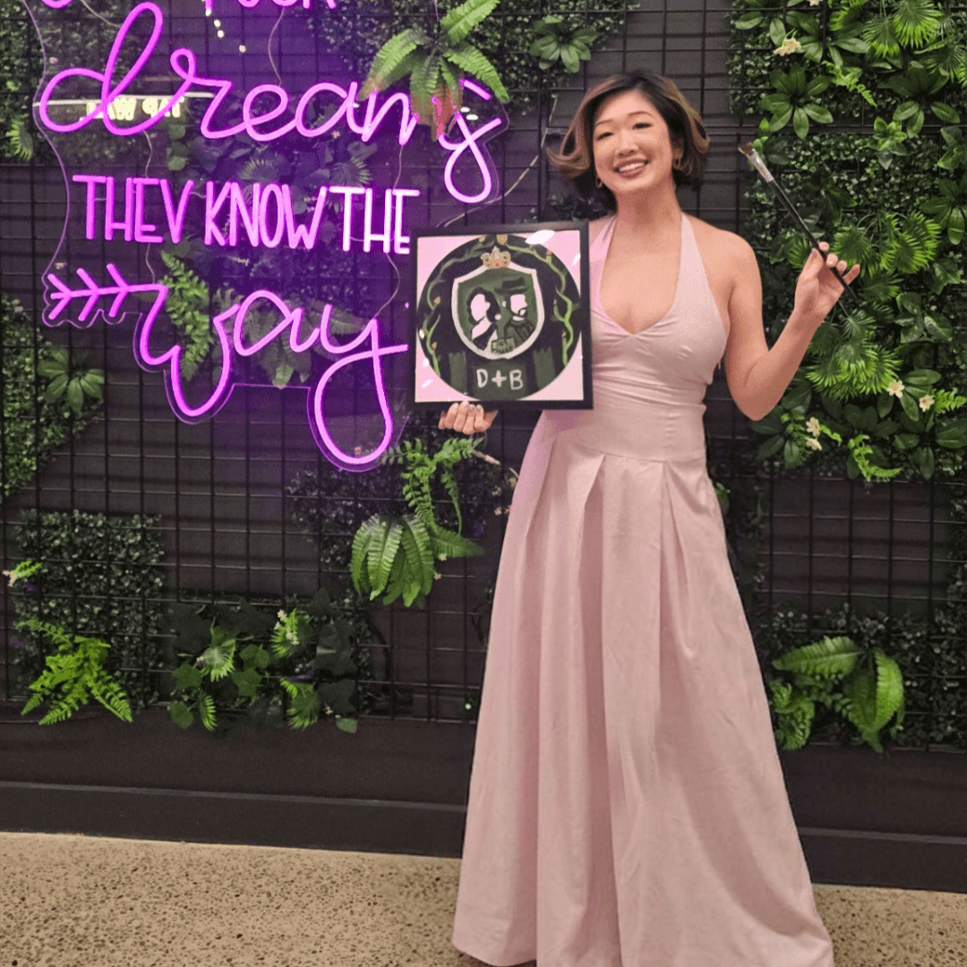 A smiling woman  in a pink dress holding a paintbrush and a finished James Bond themed painted vinyl record at Paint Cabin Toronto. Betty 