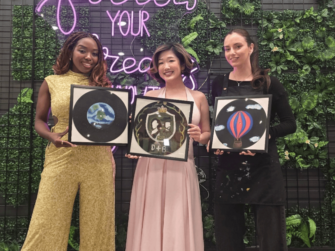 Three friends posing in a photo booth at Paint Cabin in Toronto, smiling and holding a custom painted vintage vinyl record.