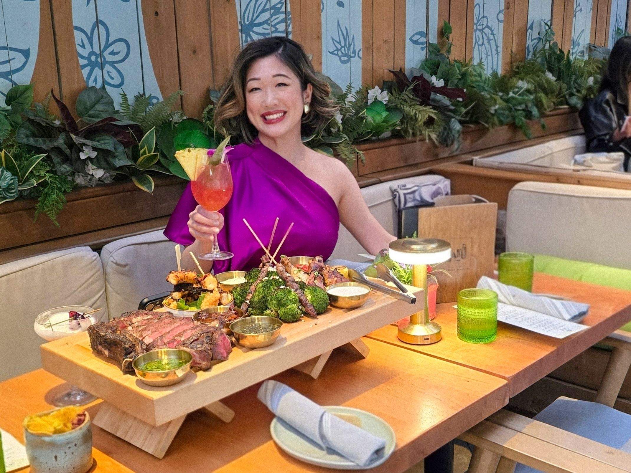 Smiling woman in a purple dress holds a cocktail at a long table with a massive steak and seafood shared board.