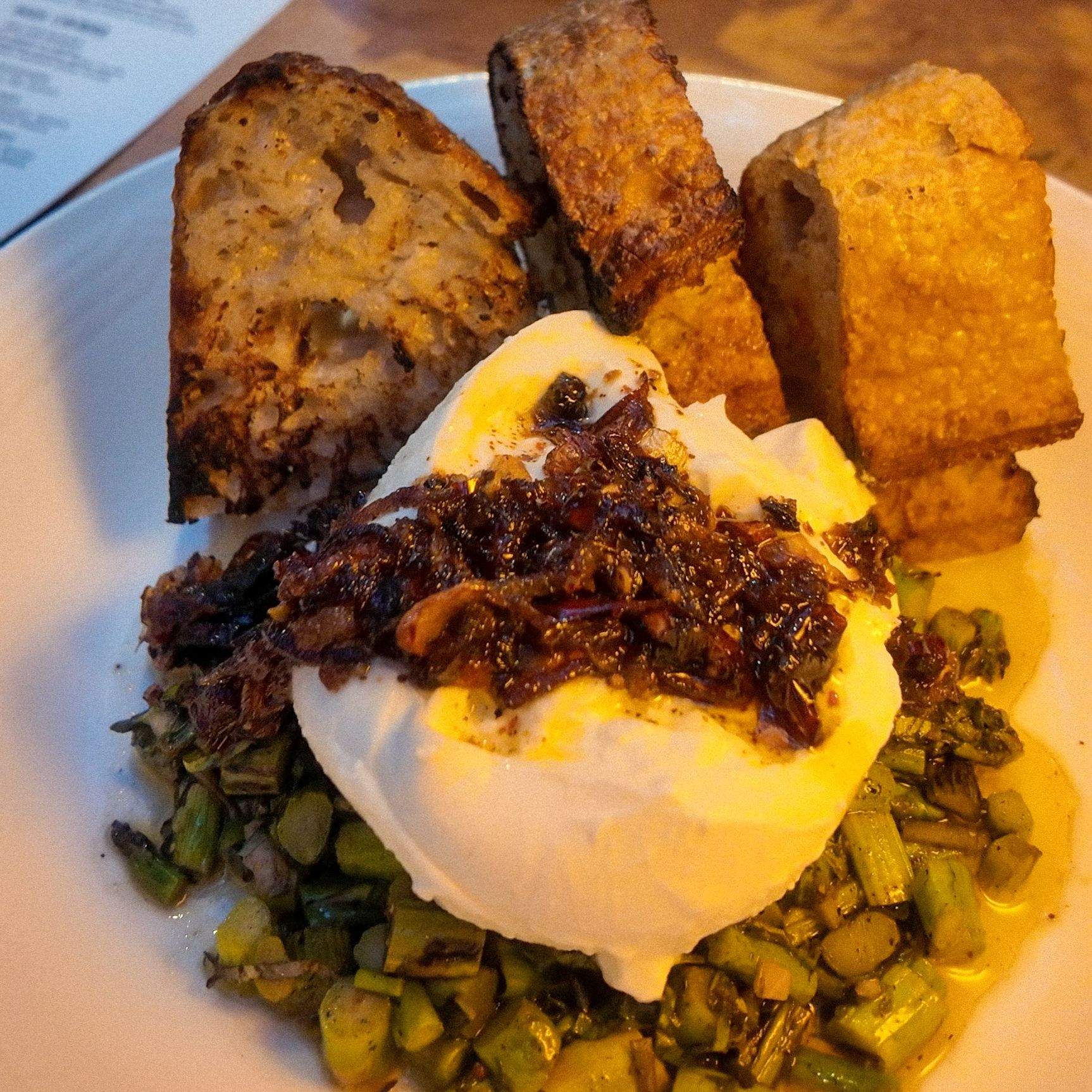 A plated appetizer of burrata cheese with garlic chili crisp, on charred asparagus with toasted sourdough.