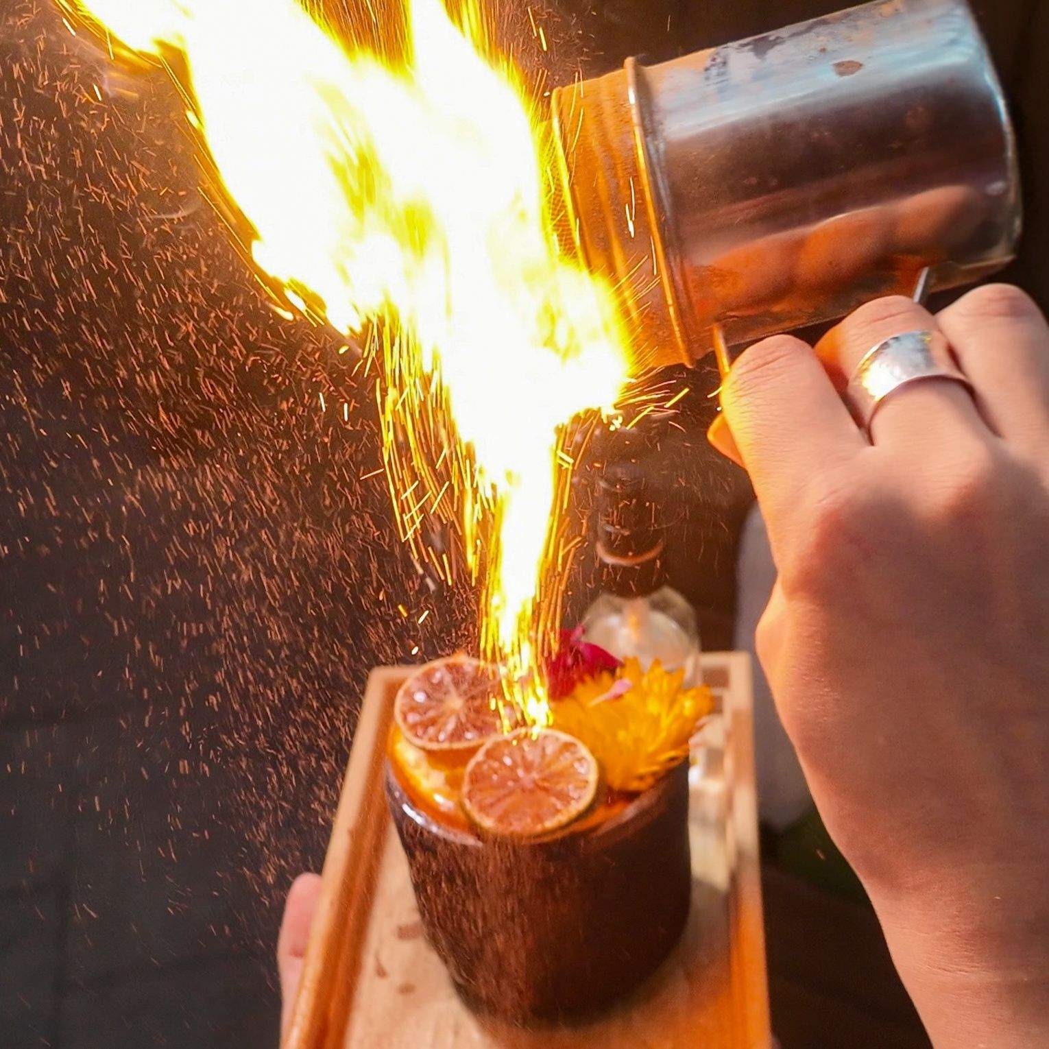 Tableside view of a server torching a flaming cocktail, with sparks, dried citrus, and flowers.