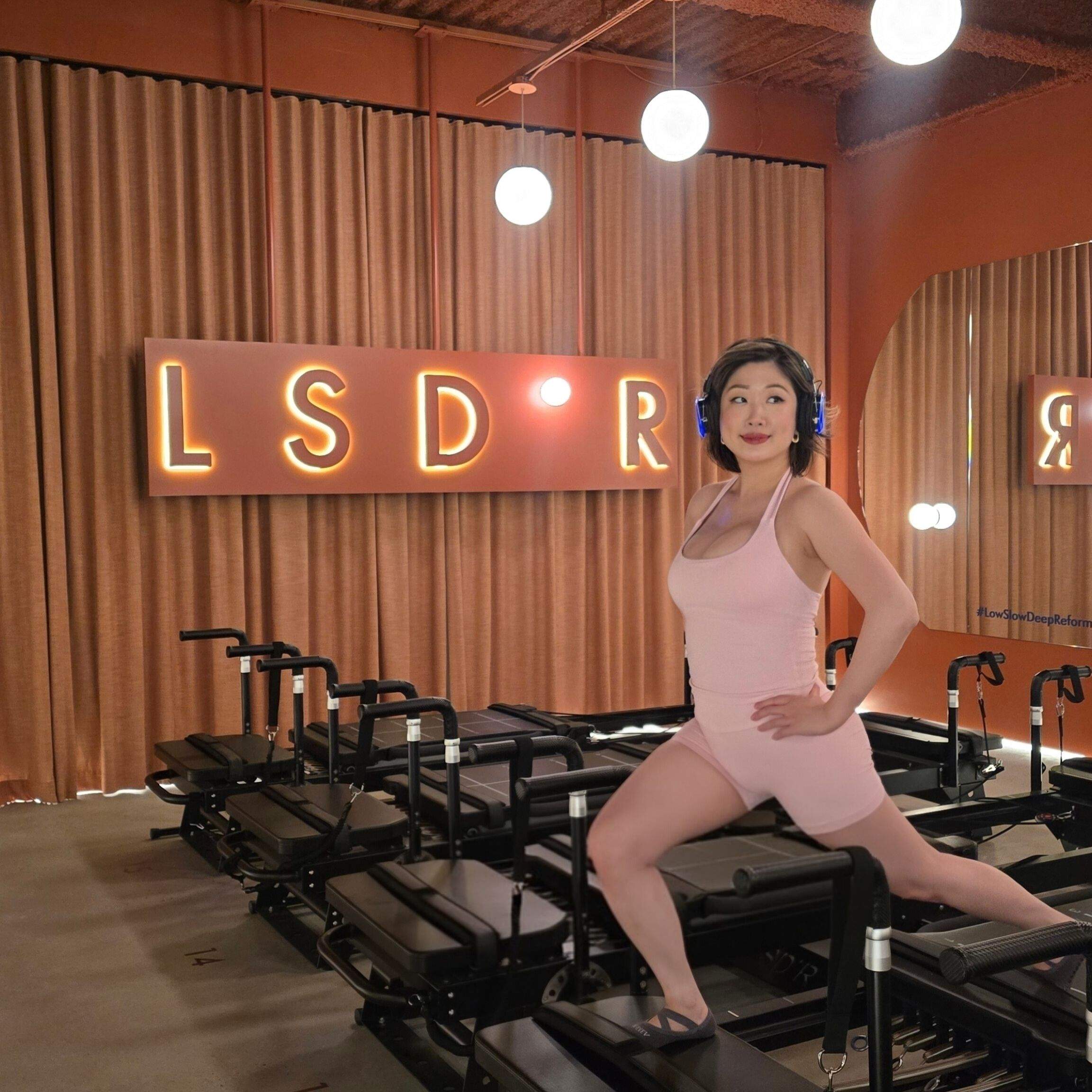 Betty posing on reformer Pilates machine at LSD°R Summerhill 1073 Yonge Street Toronto in pink activewear headphones on