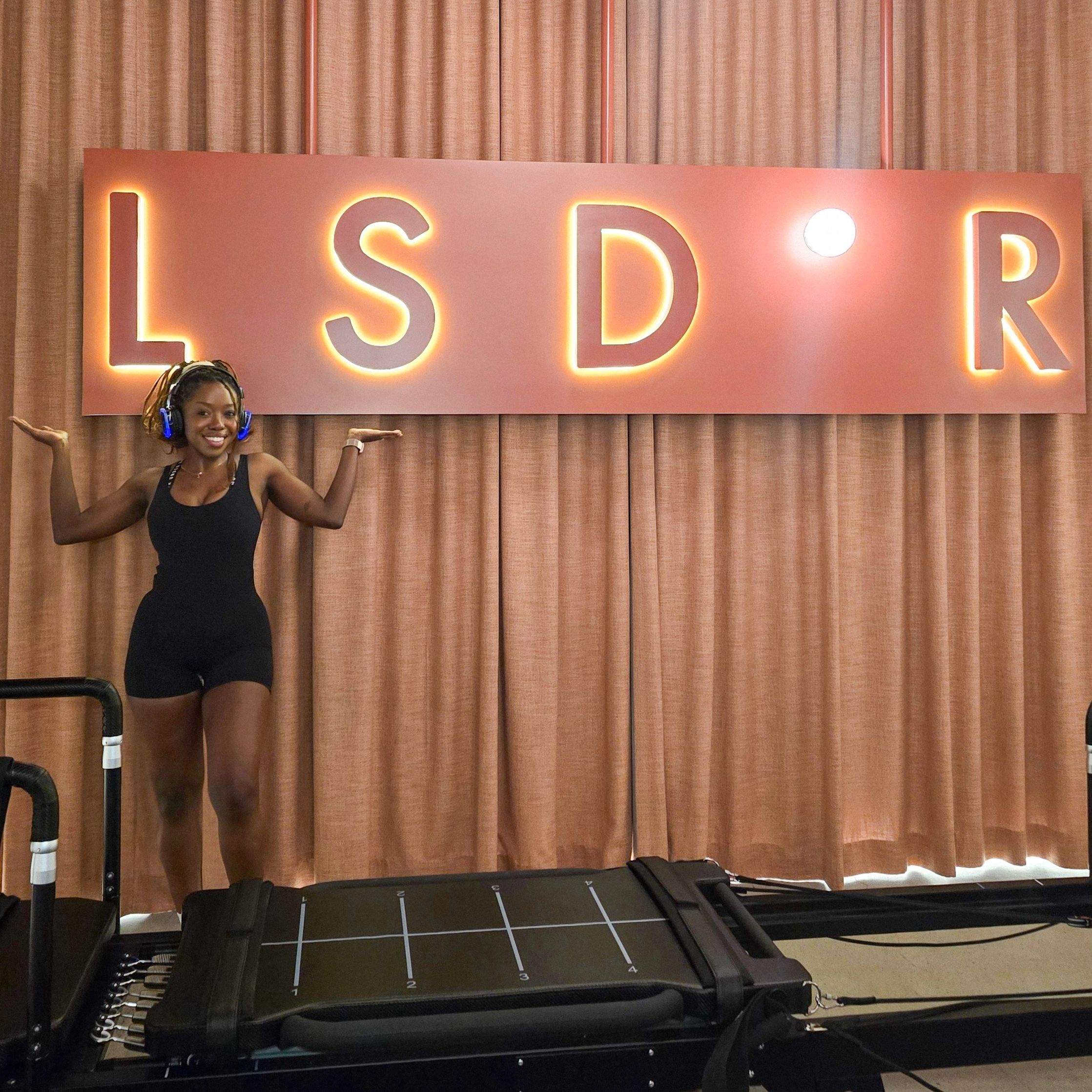 Woman posing in front of illuminated LSDR sign at LSD°R Studios Toronto boutique reformer Pilates and breathwork studio