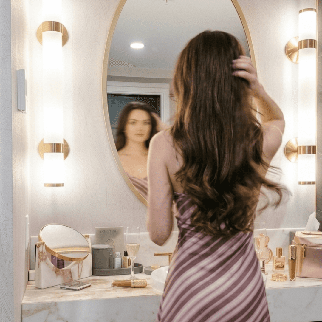Woman in a striped dress fixing her hair in front of an oval mirror in a bright marble bathroom at The Sutton Place Hotel Halifax.