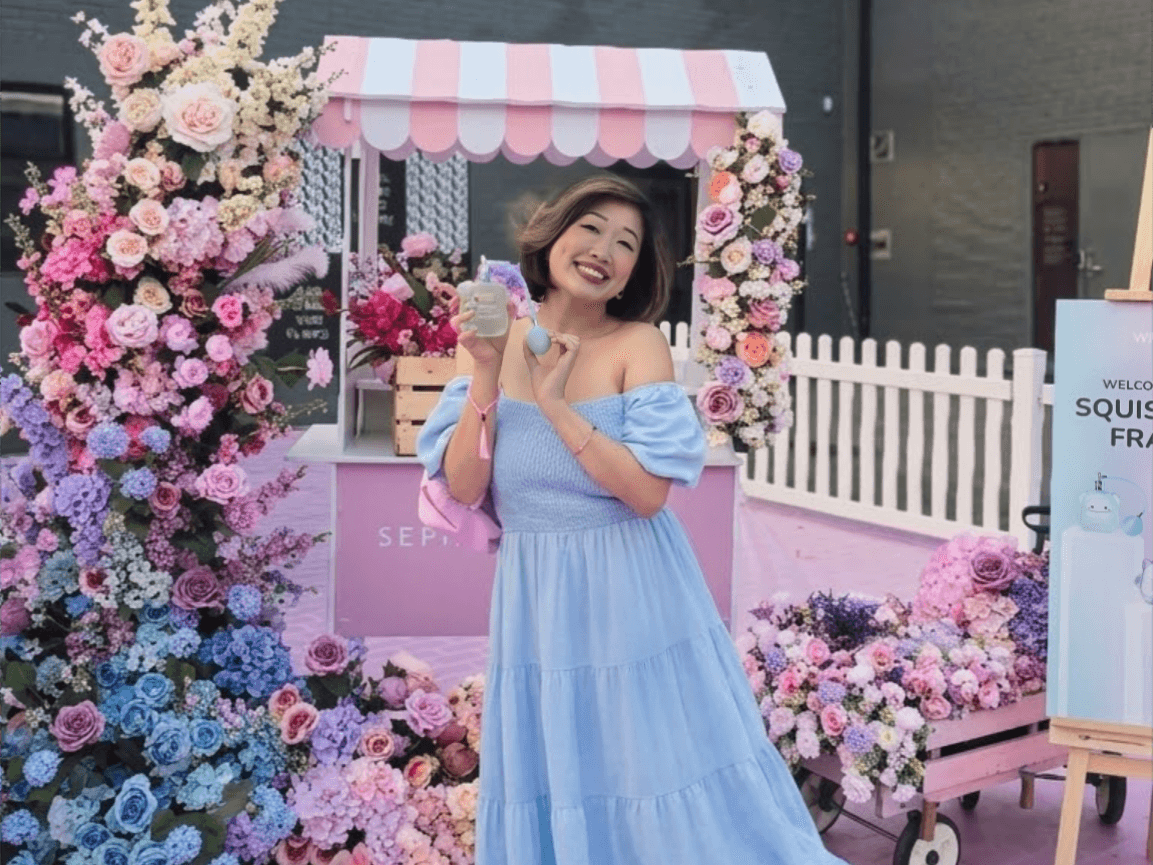 Woman in a blue off shoulder dress smiling and holding a Squishmallows Fragrances bottle in front of a pastel floral market stall.