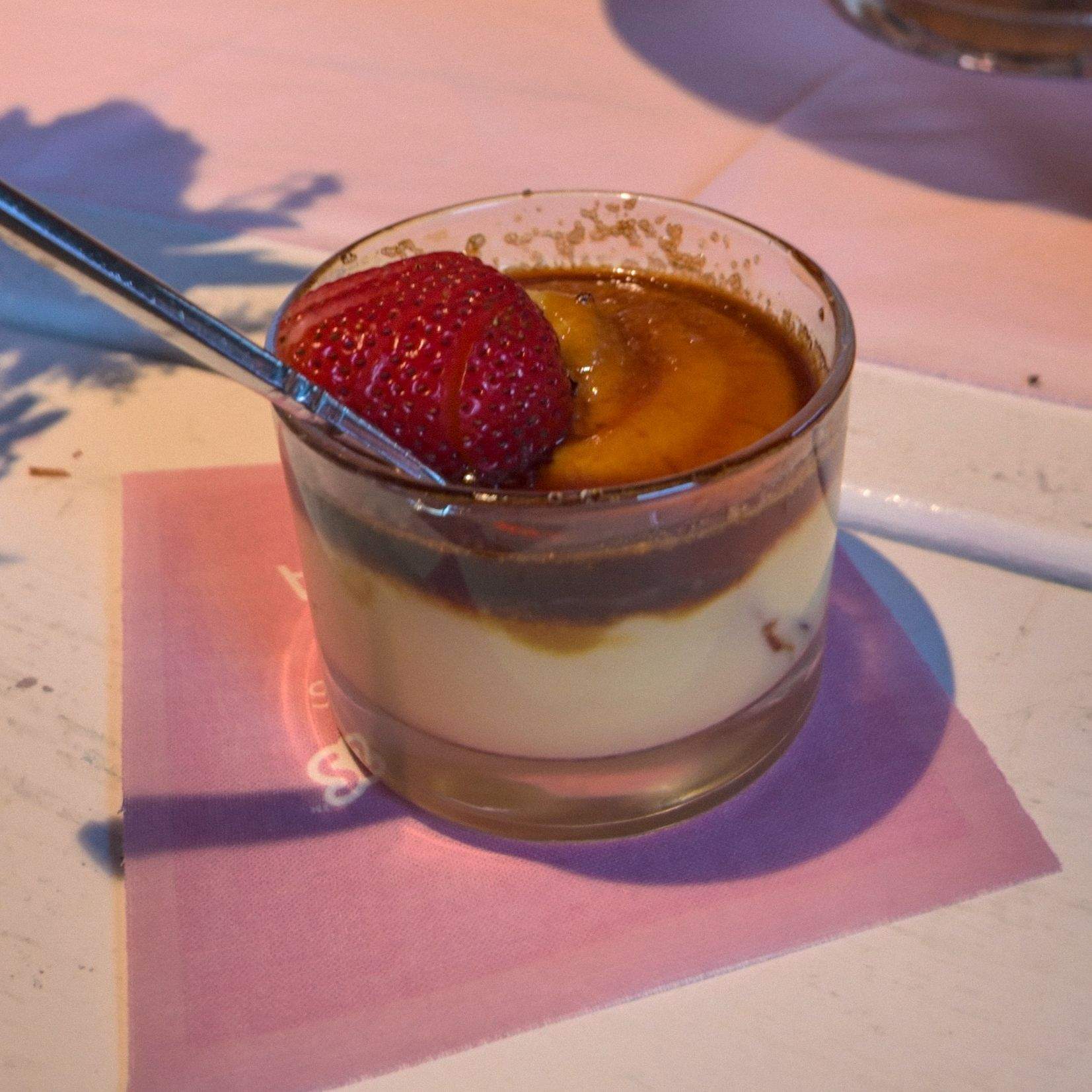 Close up of a creme brulee dessert cup with a fresh strawberry on top, cookies and another dessert cup visible behind it.