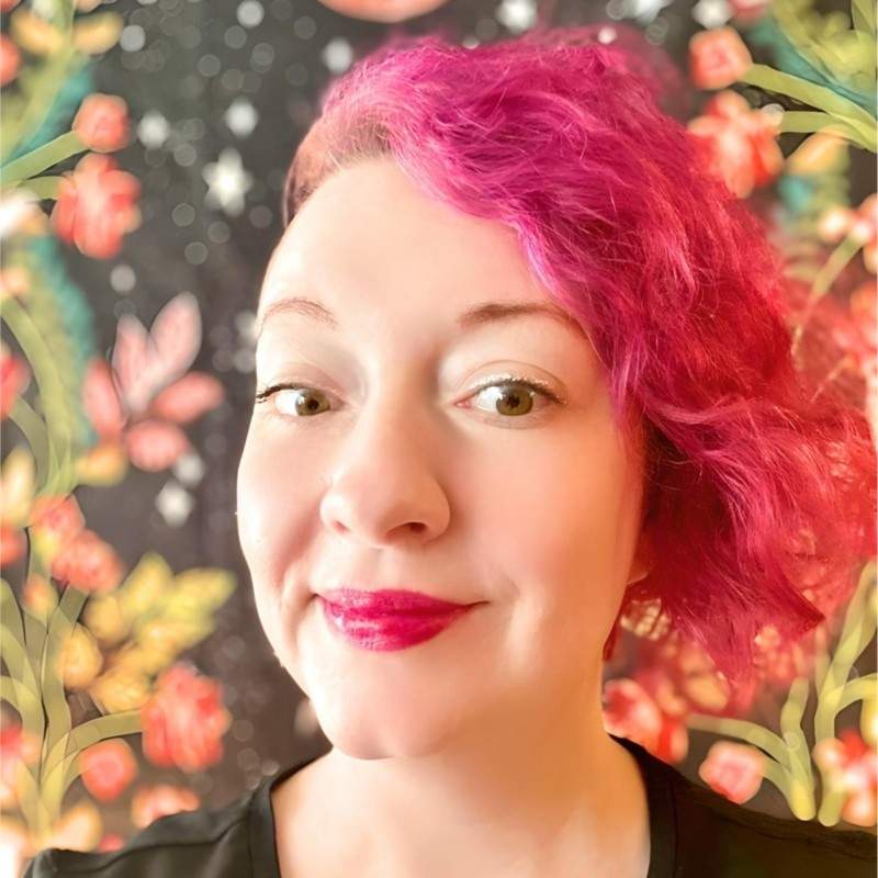 a light-skinned woman with short, curly, dark pink hair that's cropped on one side, big brown eyes and eyeliner, smiling with pink lips