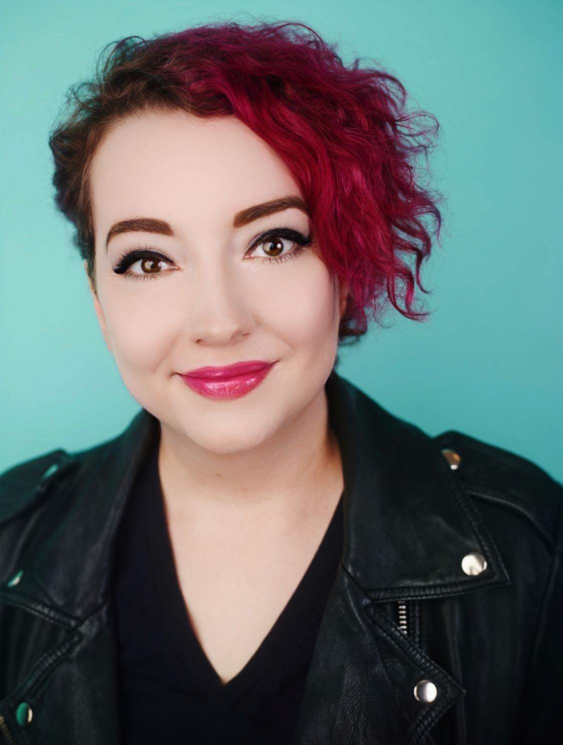 a light-skinned woman with short, curly, dark pink hair that's cropped on one side, big brown eyes and eyeliner, smiling with pink lips