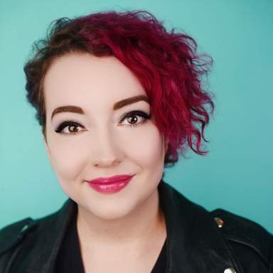 a light-skinned woman with short, curly, dark pink hair that's cropped on one side, big brown eyes and eyeliner, smiling with pink lips
