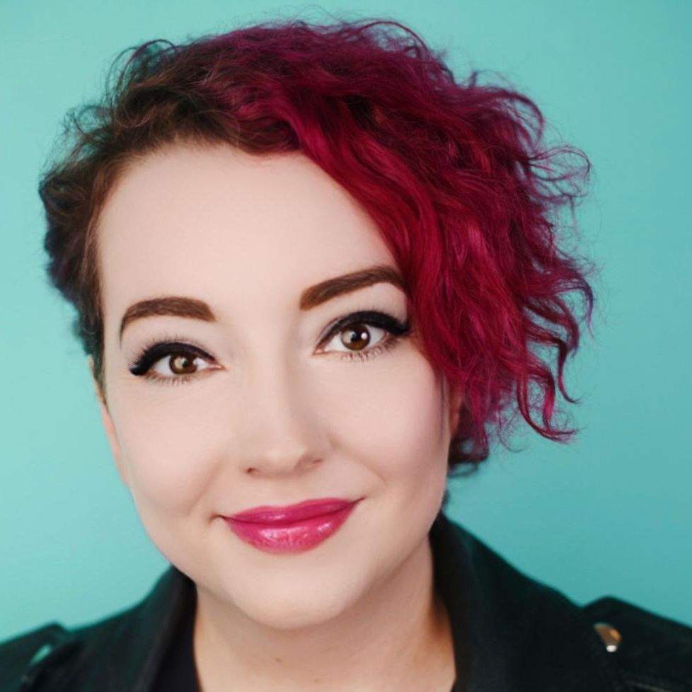 a light-skinned woman with short, curly, dark pink hair that's cropped on one side, big brown eyes and eyeliner, smiling with pink lips